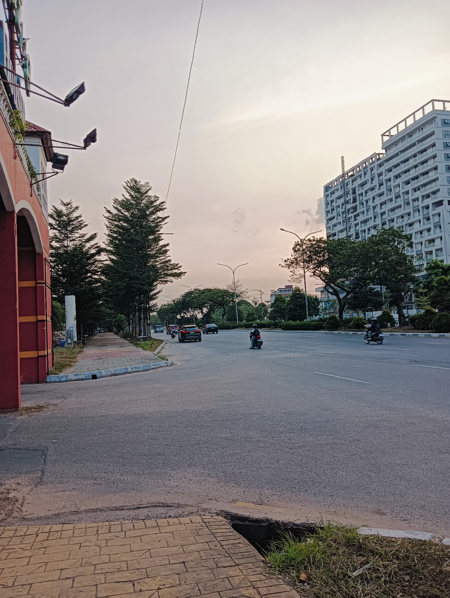 The image shows a wide, paved road in Batam, Indonesia, with a sidewalk on the left lined with trees. A large, modern apartment building is visible on the right side of the road, and another smaller building is partially obscured by trees. The sky is overcast with a pale, hazy light, suggesting either early morning or late afternoon.
Several vehicles are present on the road, including cars and motorcycles. Two motorcycles are clearly visible, one in the middle lane and another further down on the right. A dark-colored car is in the left lane. The sidewalk features a patterned paving stone surface and is bordered by a curb.
On the left side of the image, a red architectural structure with yellow accents is visible, possibly part of a building entrance. Several outdoor lights are mounted on this structure. Further down the sidewalk, a tall, white cylindrical object, possibly a utility pole, stands amongst the trees.
The overall scene depicts a typical urban streetscape in Batam, characterized by a mix of modern architecture, lush greenery, and active road traffic.