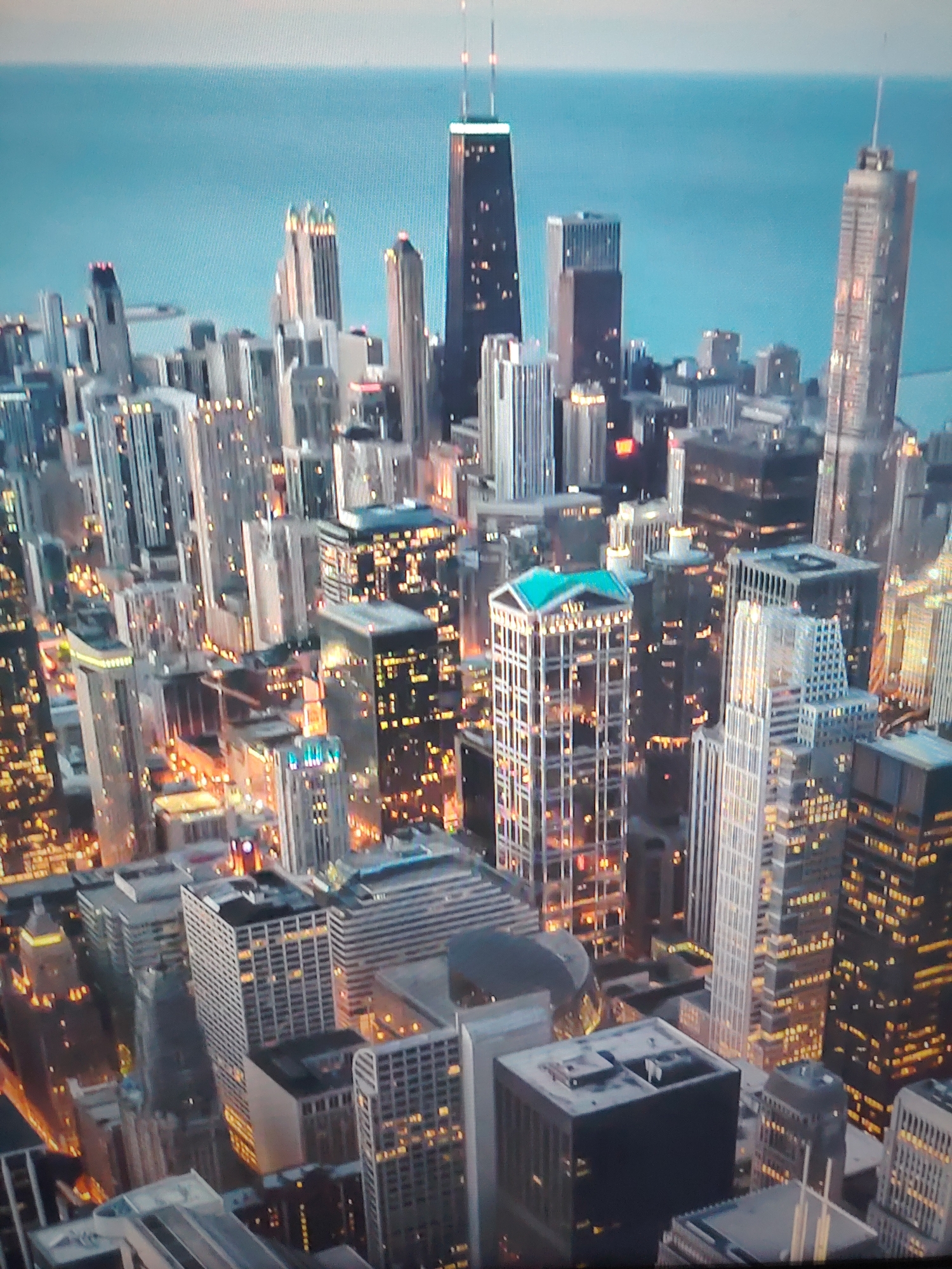 The image depicts a high-angle view of a dense urban cityscape at dusk or early evening. The location is Chicago. A multitude of skyscrapers dominate the skyline, many illuminated with artificial lighting. The John Hancock Center is prominently visible, and the Willis Tower is also visible. A large body of water appears in the background. Streets are illuminated with orange-yellow lights. The environment suggests a bustling metropolis at a time when the transition from daylight to night is occurring.