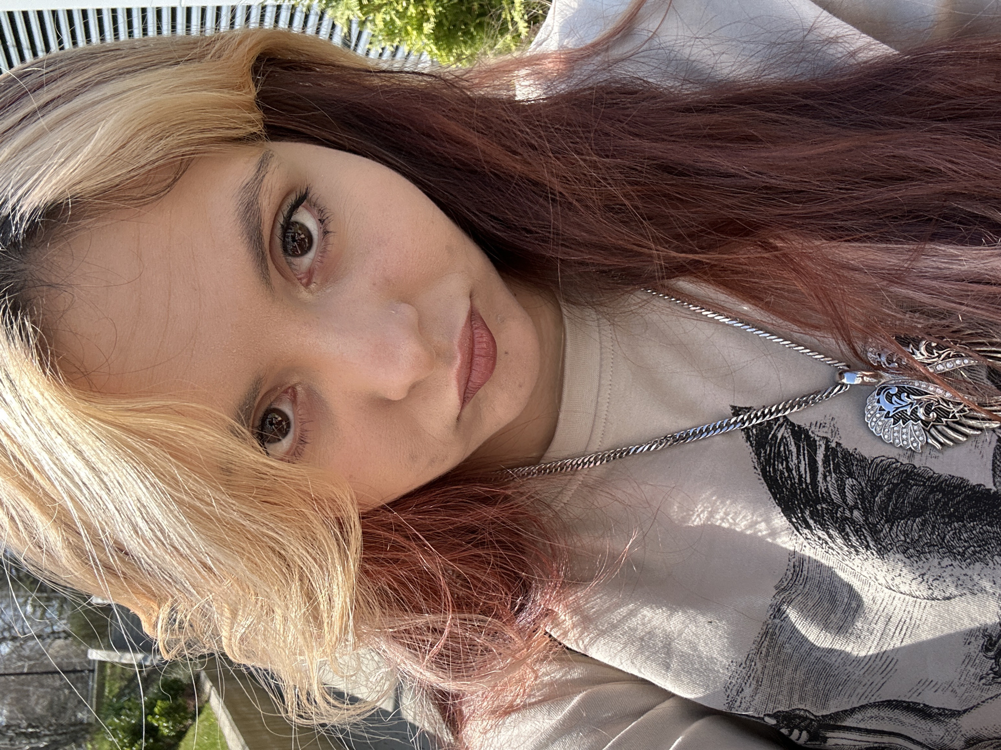 A person is captured in a close-up shot, tilted slightly, with their gaze directed at the camera. They have a fair complexion, brown eyes, and multicolored hair featuring blonde streaks on one side and auburn shades on the other. The individual wears a light-colored sweatshirt imprinted with a darker graphic design. A silver chain necklace with a pendant adorns their neck. The background shows glimpses of greenery and a barred fence under bright sunlight. There is no explicit indication that the location is Sofia, Bulgaria.