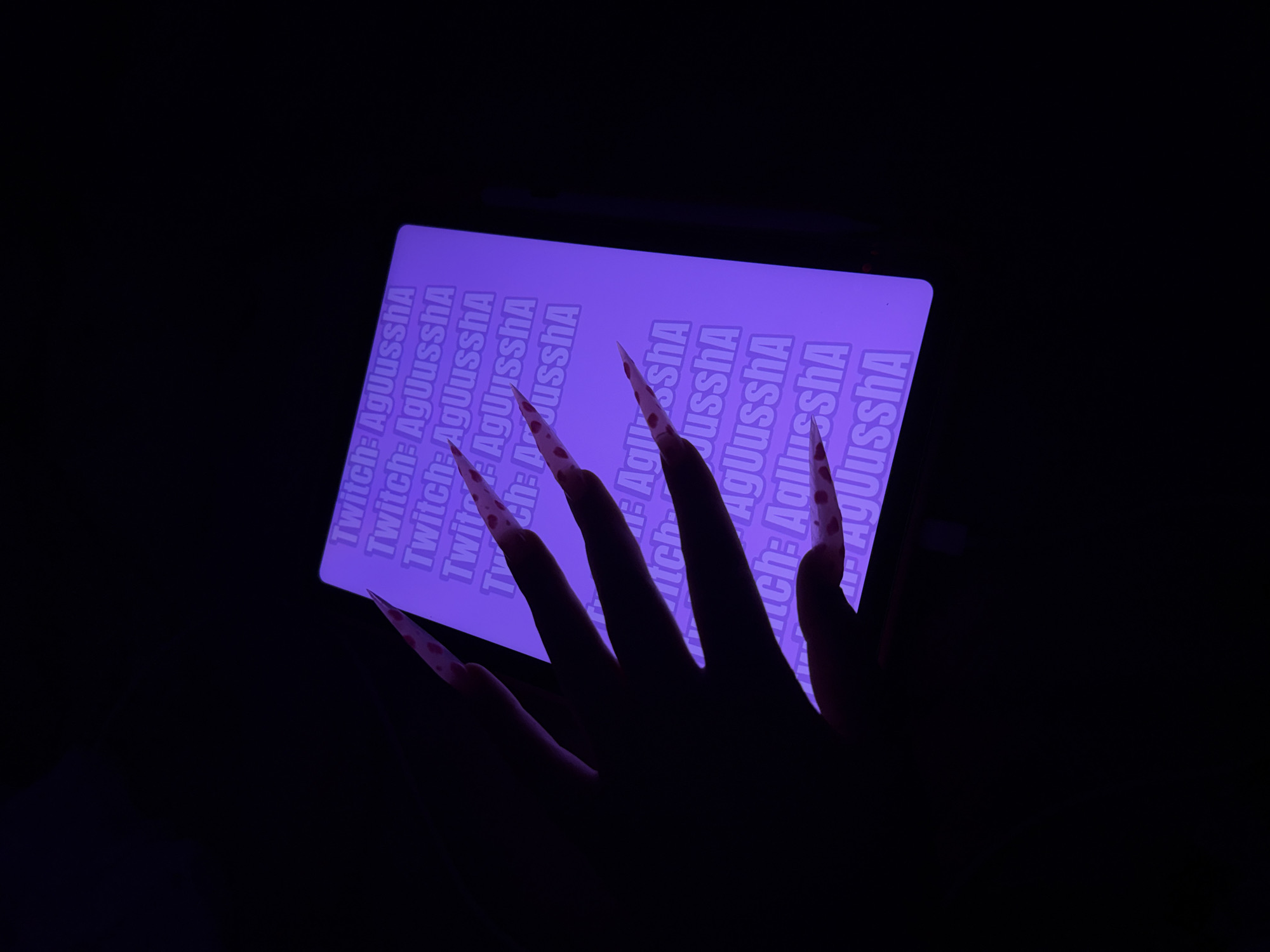 A hand with long, pink, heart-patterned acrylic nails is held up in front of a dark background. The hand is partially silhouetted against a bright purple screen displaying repeated text that reads "Twitch: AgUusshA". The screen appears to be a tablet or monitor, and the text is white with a slight purple hue. The scene is dimly lit, with the purple screen being the primary light source. The location of Pudomyagi, Russia is not evident in the image.