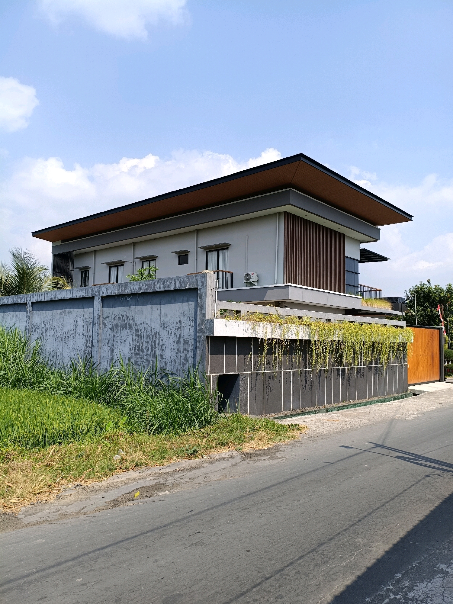 The image shows a modern, two-story house with a flat roof, situated in Purwomartani, Indonesia. The house is painted light gray and features dark wooden accents on its exterior. A prominent, high concrete wall surrounds the property, with lush green grass and some dry foliage growing along its base. The wall is textured and shows signs of weathering. Behind the wall, the house's upper stories are visible, with windows and balconies. A section of the wall near the right side of the image has a wooden gate. The foreground displays a paved road with some cracks and a shadow cast by an unseen object, likely a lamppost. The sky above is a clear blue with scattered white clouds.