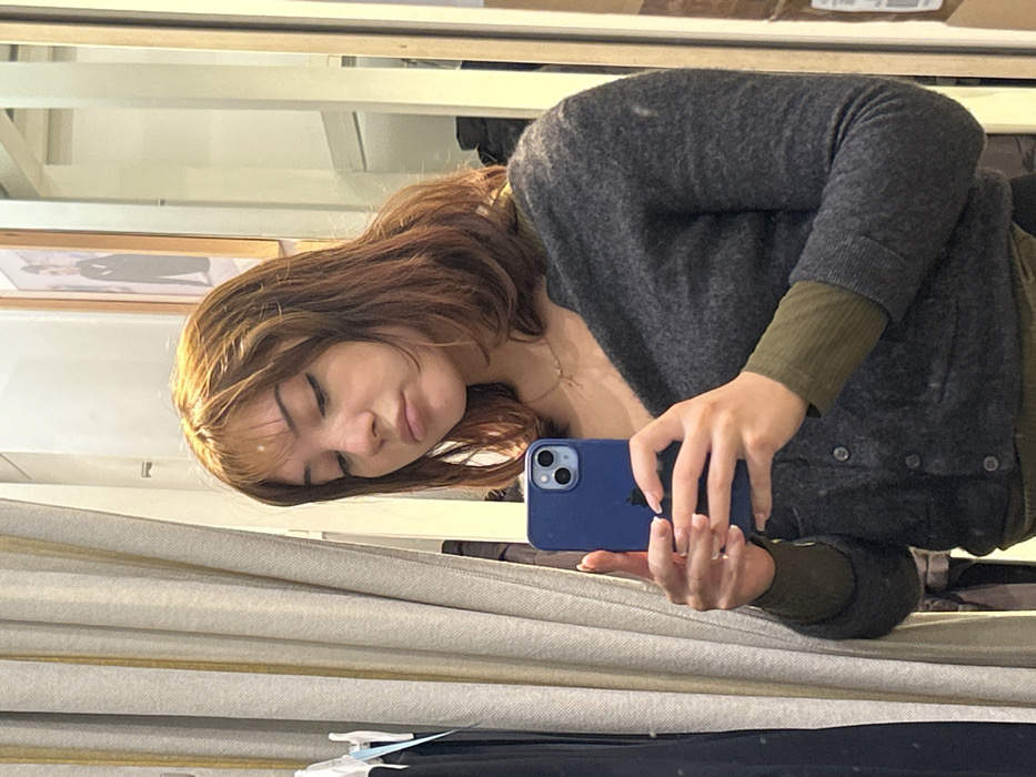 A young adult female is depicted taking a mirror selfie indoors, capturing her reflection from the mid-chest upward. She possesses shoulder-length, layered reddish-brown hair with lighter bangs and face-framing highlights. Her expression is a slight pout, with her eyes directed downward toward the smartphone. She is dressed in a dark gray button-up cardigan layered over an olive green long-sleeved shirt, complemented by a delicate gold-toned necklace. Both hands grasp a blue-cased smartphone, prominently displaying its dual camera system. The background to the left features a light-colored, vertically striped curtain partially obscuring a rack with dark garments. To the right, a light-colored wall with a framed picture is visible, along with vertical reflective surfaces or partitions, suggesting a retail fitting room or similar indoor commercial space. The event occurs in the city of Strasbourg, France.