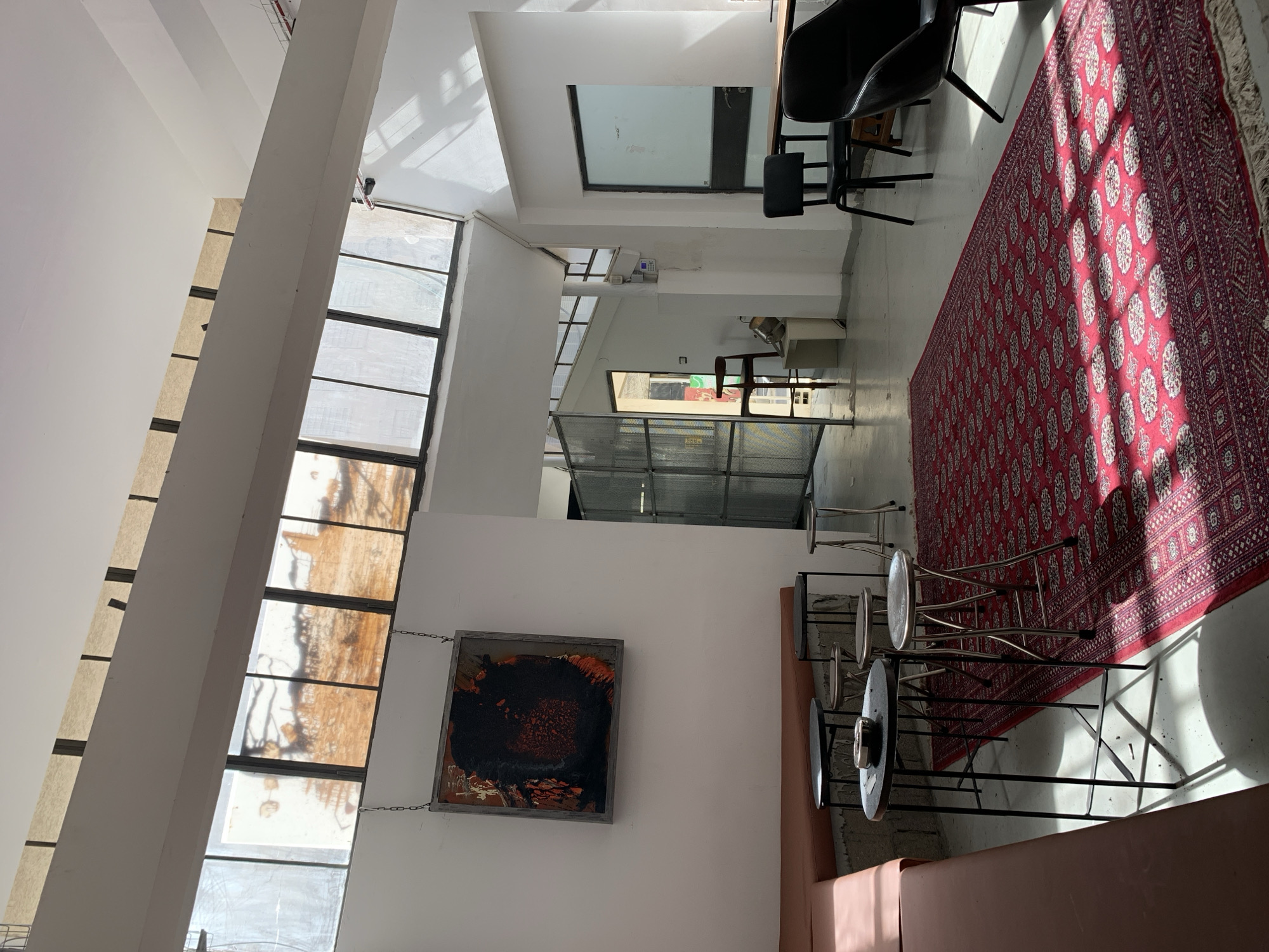 This is an interior shot in Tel-Aviv, Israel, featuring a room with a mix of modern and rustic elements. Sunlight pours through a tall, multi-paneled window on the left, illuminating a section of the room. A dark-framed abstract artwork hangs on a white wall near the window. Several metal-framed stools are arranged near a leather-like banquette seating area. A large, patterned red rug dominates the right side of the room. In the background, a glass partition divides the space, revealing another area with furniture and glimpses of the outside. The overall environment has a contemporary, loft-like feel.