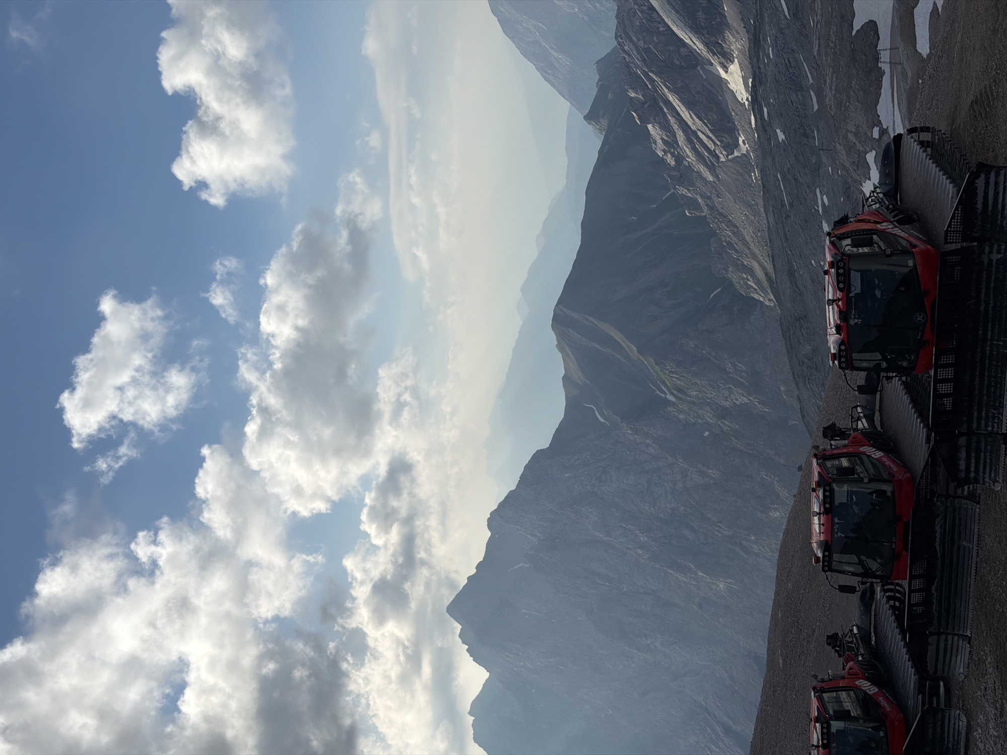 The image captures a scenic mountain landscape near Garmisch-Partenkirchen, Germany, under a bright blue sky dotted with white, fluffy clouds. In the foreground, three red snow groomers are parked on a gravelly surface. The machines have a distinctive tracked design and a cab with windows. Behind the snow groomers, a vast mountain range stretches into the distance, with peaks becoming progressively fainter and bluer due to atmospheric perspective. Some snow is visible on the higher slopes, suggesting a high-altitude environment. The overall scene is one of natural grandeur combined with the presence of specialized vehicles.
