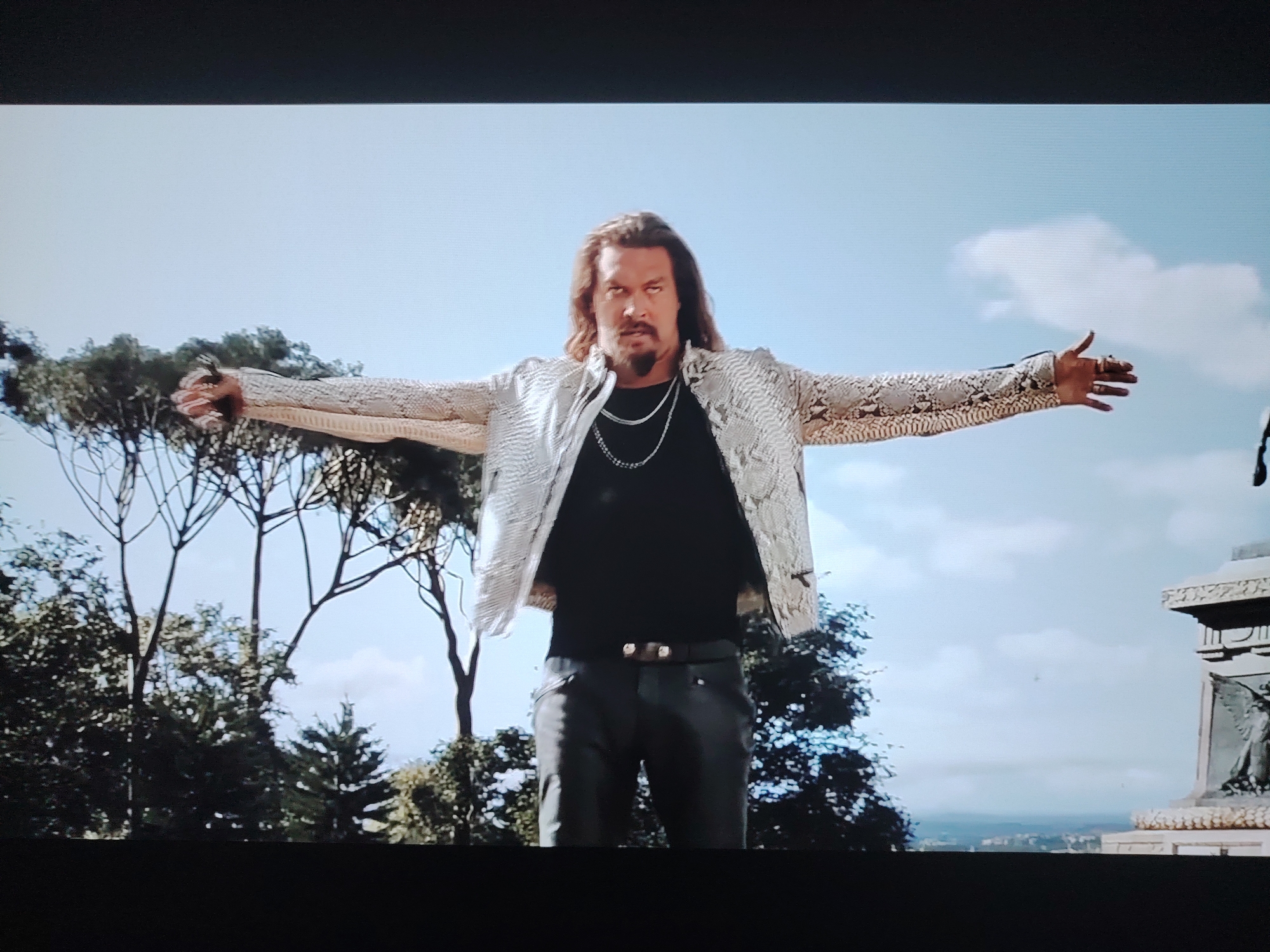 A man with long, brown hair and a goatee stands outdoors with his arms outstretched. He is wearing a black t-shirt, a snakeskin-patterned jacket, and dark pants with a brown belt. The man is looking directly at the camera with a serious expression. He is wearing two silver necklaces and several rings on his fingers. In the background, there are trees, a blue sky with clouds, and what appears to be a monument. The scene is likely outdoors in a city, but the specific location in Huntingdonshire, United Kingdom, cannot be determined from the image.