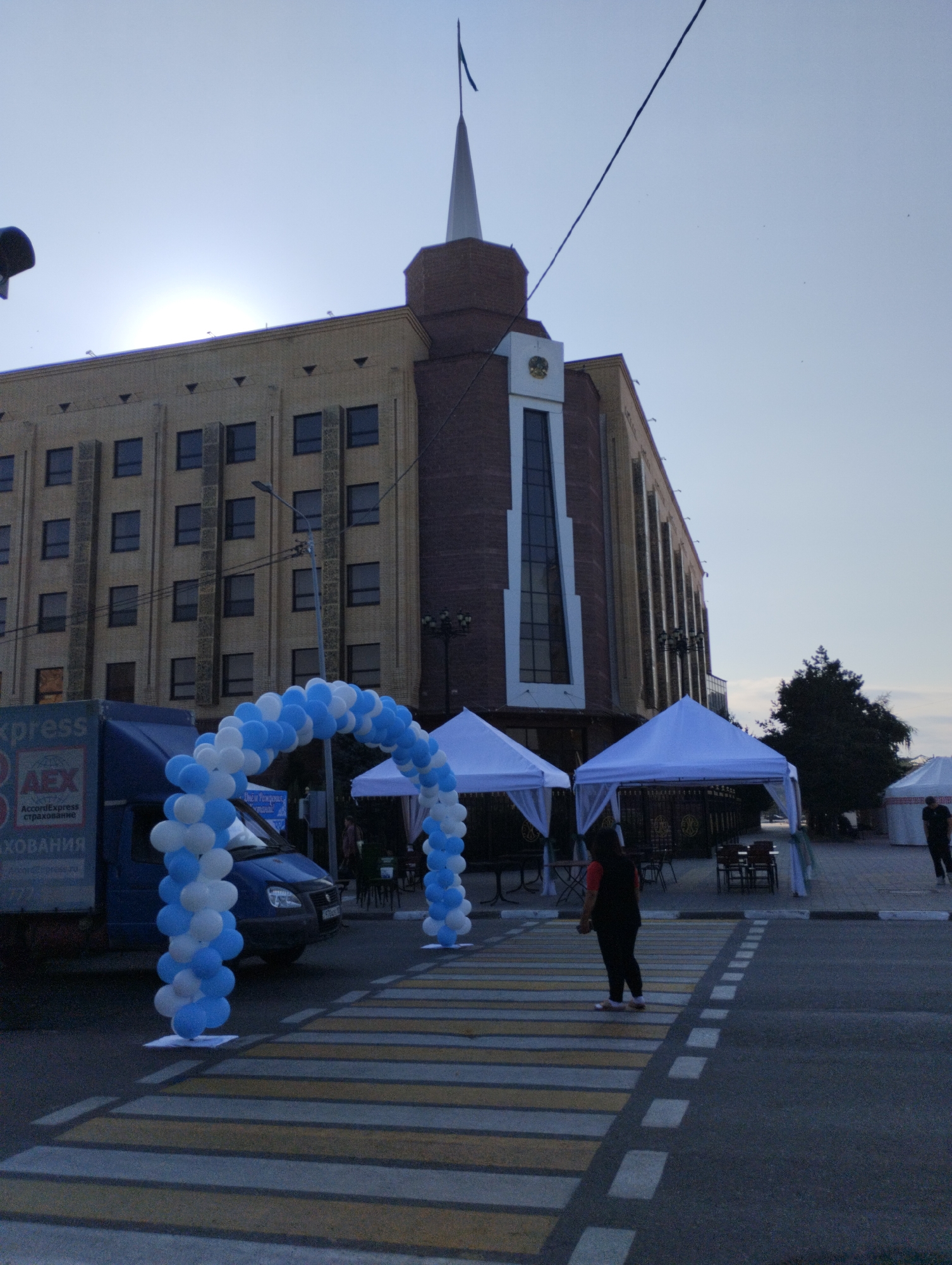 A wide-angle, low-light, outdoor photograph captures a street scene in front of a large, modern building. The building, with a cream-colored facade and dark brick accents, features multiple rows of rectangular windows. A tall spire with a flag tops a section of the building. In the foreground, a blue truck with text in Russian, including "xpress" and "AEX AccordExpress страхование", is parked on the left. A pedestrian crosswalk, marked with yellow and white stripes, is prominent. Two arched balloon decorations, one blue and white, the other primarily blue, span across the street. Several white tents are set up, and a person wearing a red shirt and black pants stands on the crosswalk facing away from the camera. Another person in dark clothing is visible in the distance to the right. The sky is a pale blue, suggesting either early morning or late evening light. The overall impression is of an event or market setup on a city street.