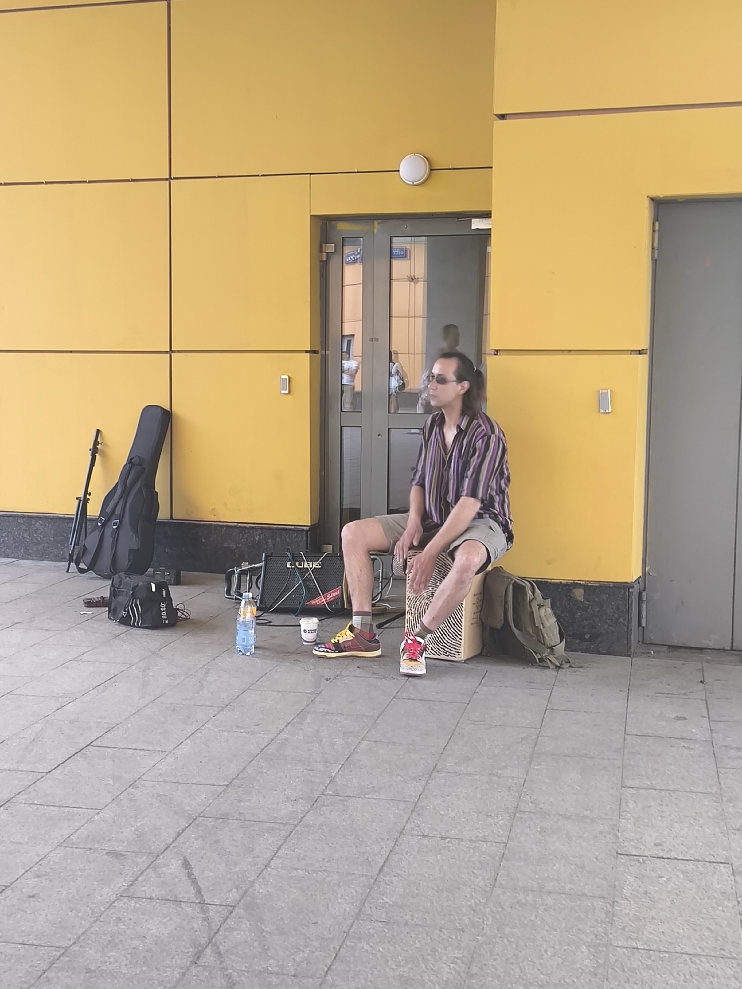 A street musician sits outside a yellow building with a gray doorway, playing a box drum. He wears a striped shirt, khaki shorts, and colorful sneakers. A guitar in a black case and a small amplifier are nearby, along with a water bottle and a backpack. The location appears to be an urban public space with a tiled floor.