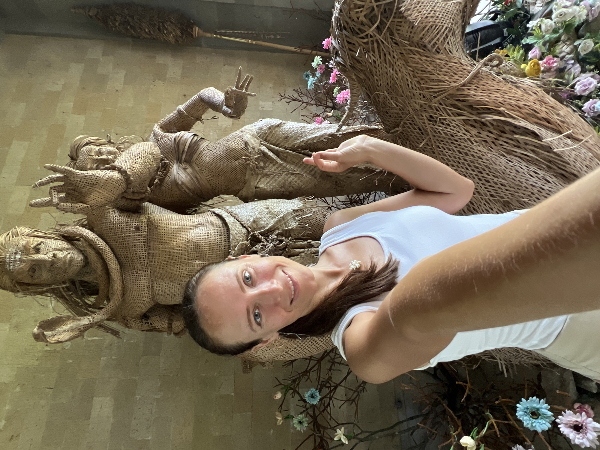 The image is a selfie taken indoors in Kedewatan, Indonesia. A woman in a white tank top smiles at the camera. Behind her are two reclining figures made of woven material, resembling statues of deities. One is lying flat and the other on an upper level. There is a display of artificial flowers and dried plant material. The wall is a light tan color.
