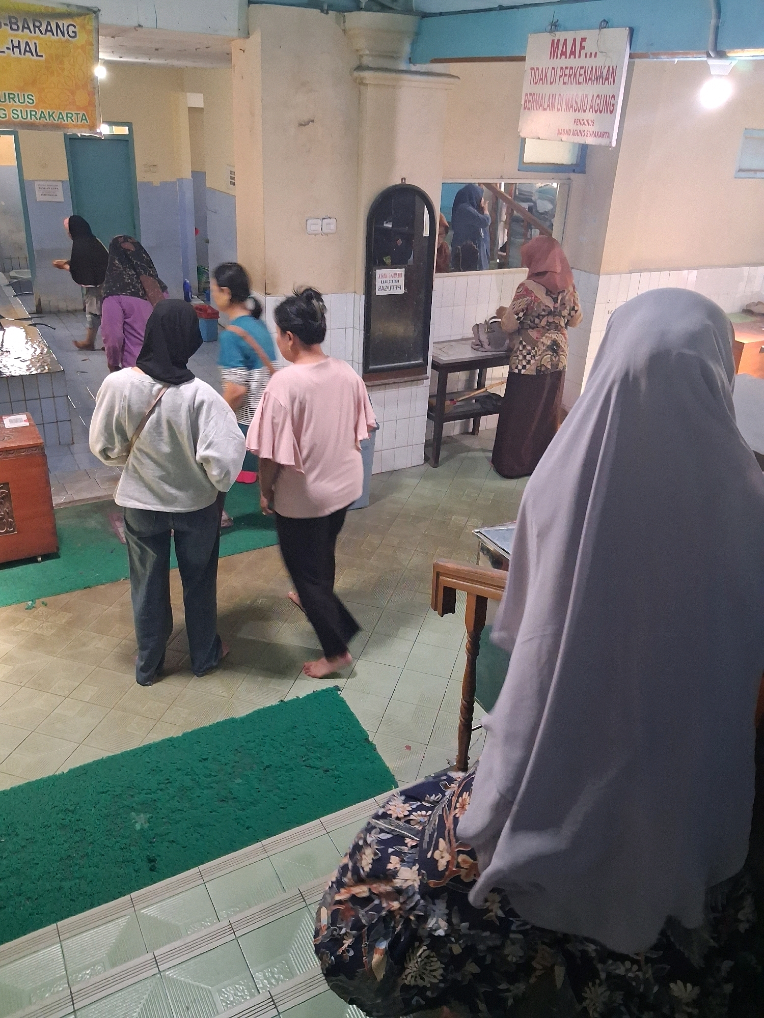The image depicts an indoor scene, likely a public building in Surakarta, Indonesia, given the presence of Indonesian text on signage. In the foreground, a person wearing a grey hijab and a floral patterned garment is seated, with their back to the camera. Their face is not visible. To their left, a person wearing a light grey long-sleeved shirt and dark pants stands with their back to the camera, looking towards the left. To the right of this person, another individual in a light pink shirt and dark pants walks away from the camera, also barefoot.
In the mid-ground, several other individuals are visible. One person in a patterned top and a dark skirt is standing near a wooden table and chairs. Another person in a blue shirt is walking away from the camera. In the background, a person wearing a black hijab and a purple garment is visible near a counter.
Various objects are present in the scene, including a wooden chest on the left, a wooden table with chairs, and a green mat on the floor. Signage is visible on the wall. One sign, in Indonesian, reads "MAAF... TIDAK DI PERKENANKAN BERMALAM DI MASJID AGUNG PENGURUS MASJID AGUNG SURAKARTA," which translates to "SORRY... OVERNIGHT STAY IS NOT ALLOWED IN THE GRAND MOSQUE, MANAGEMENT OF THE GRAND MOSQUE SURAKARTA." Another sign on the left, also in Indonesian, appears to be related to goods or services.
The environment appears to be a well-lit interior space with tiled floors and light-colored walls. The overall atmosphere suggests a public area within a mosque or a similar institution.