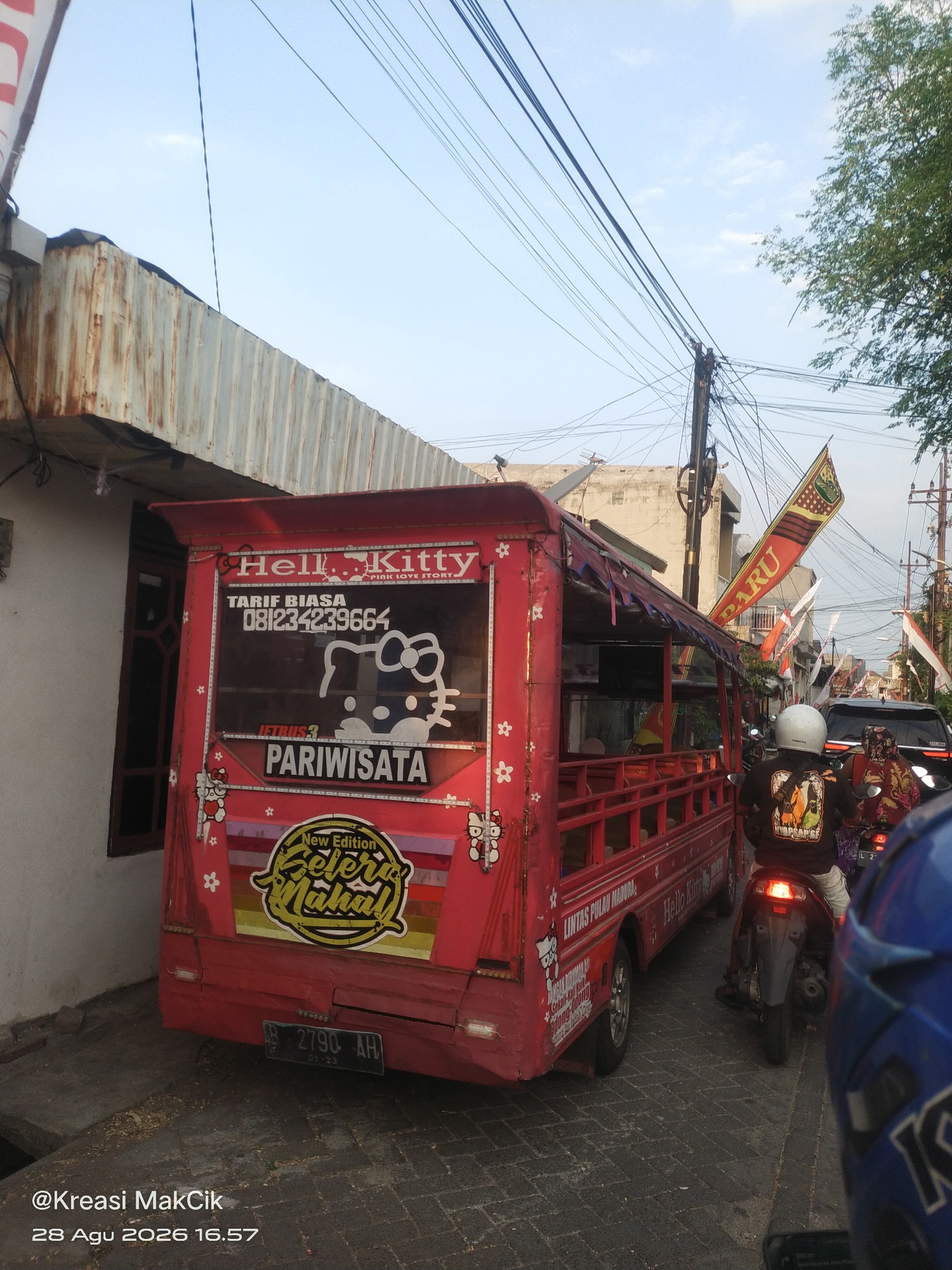 A red, open-sided vehicle, possibly a type of jitney or shuttle bus, is parked on a cobblestone street in Surabaya, Indonesia. The vehicle is adorned with Hello Kitty imagery and features text in Indonesian, including "TARIF BIASA," "PARIWISATA," and "New Edition Selera Mahal." A license plate with the markings "B 2790 AH" is visible on the rear.
The street is lined with buildings and utility poles with numerous overhead wires. Red and white Indonesian flags are displayed on poles along the street, suggesting a possible local celebration or event.
In the foreground and background, several motorcycles are present. One motorcycle carries two individuals, with the passenger wearing a colorful headscarf and the driver sporting a black t-shirt with a graphic design. Another motorcycle is visible further back. A black car is also in the scene, suggesting it is a relatively busy urban area. The sky is partly cloudy, and the overall lighting suggests it is daytime. The presence of the utility poles and the style of buildings indicates a Southeast Asian urban environment.