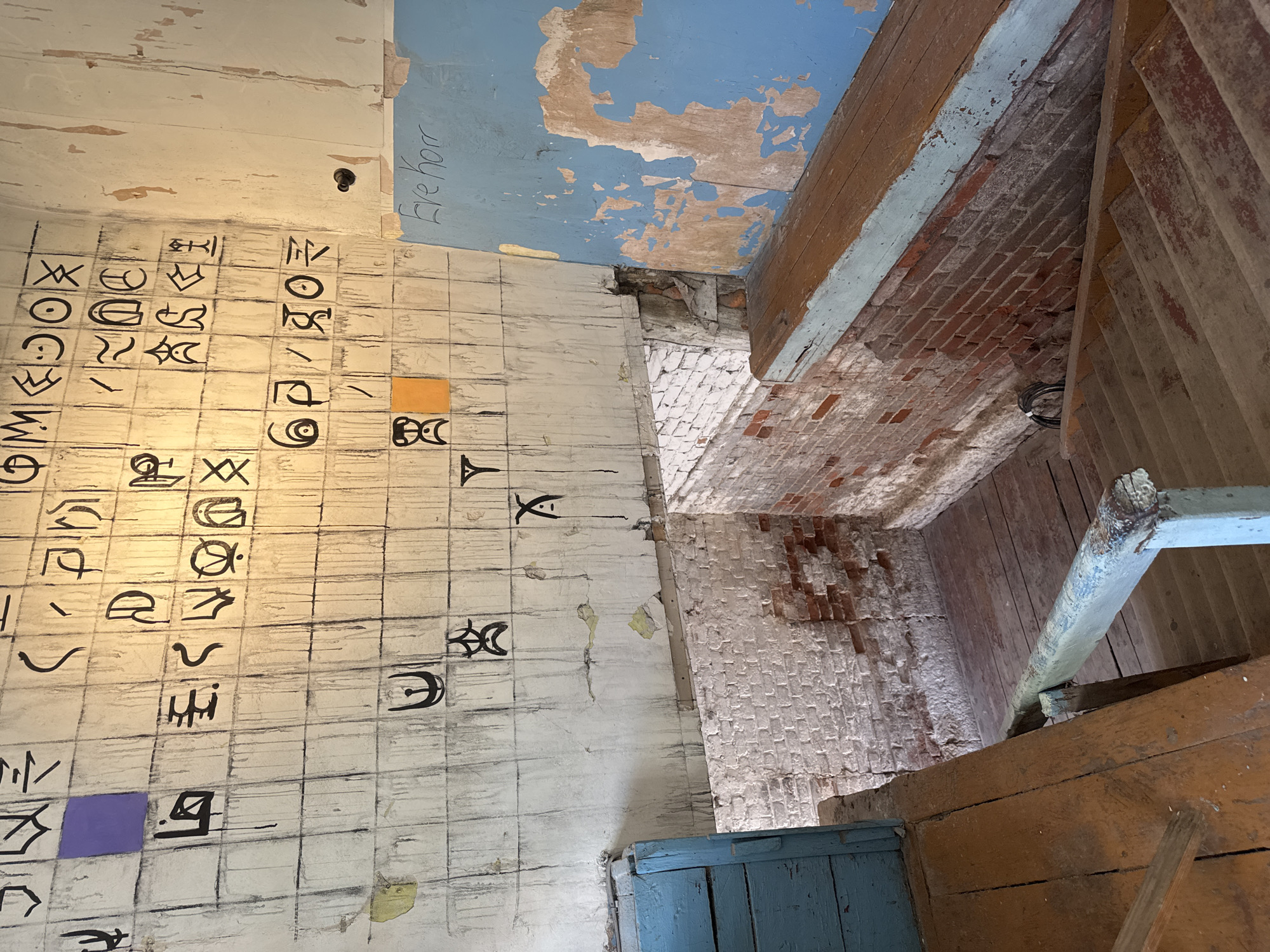 The image depicts a weathered interior space with a prominent wall covered in a grid of squares, some containing black symbols and others left blank. There are streaks of black paint running down the wall, suggesting artistic execution or deterioration. Two small squares, one purple and one orange, are affixed to the grid. To the right, a section of blue-painted wall is visible, with peeling paint revealing lighter material underneath. Handwritten text in black marker reads "Eve Horr" on this blue section.
In the foreground, wooden stairs descend into a lower level. The stairs and surrounding platforms are made of worn, unfinished wood, showing signs of age and use. A light blue wooden door is visible on the left side of the stairs. The lower level walls are constructed of exposed brick, with some sections whitewashed and others showing the raw brick. The overall impression is of an abandoned or neglected space, possibly an art installation or an old industrial building.
The location is identified as the former administrative building of the Yelets Confectionery Factory in Yelets, Russia. This building is known for its artistic transformations, including the wall art visible in the image, created by artist Valery Gryaznov.