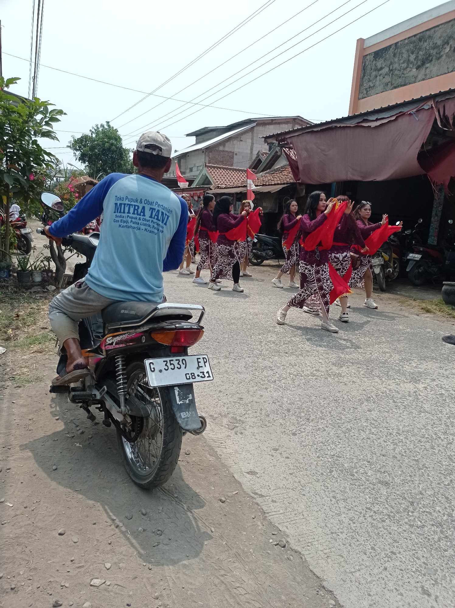 The image depicts a street scene in Kramat, Indonesia, with a group of young women performing a traditional dance. The dancers, dressed in matching purple tops and patterned skirts with red accents, are holding red cloths. They are positioned on the right side of the paved road, seemingly in motion. In the foreground, a man is seated on a motorcycle, facing away from the camera. He is wearing a light blue long-sleeved shirt with white lettering that reads "Toko Pupuk & Obat Pertanian MITRA TANI" and "Sedia: Pupuk & Obat Pertanian Gas Elpiji, Pulsa Listrik, Pulsa HP, Kertaharja - Kramat". His motorcycle has a license plate that reads "G. 3539 FP GB-31". The background shows buildings with red tile roofs and overcast sky. The environment appears to be a residential or commercial area in a rural or semi-urban setting, suggested by the presence of houses and shops. The lighting indicates it is daytime.