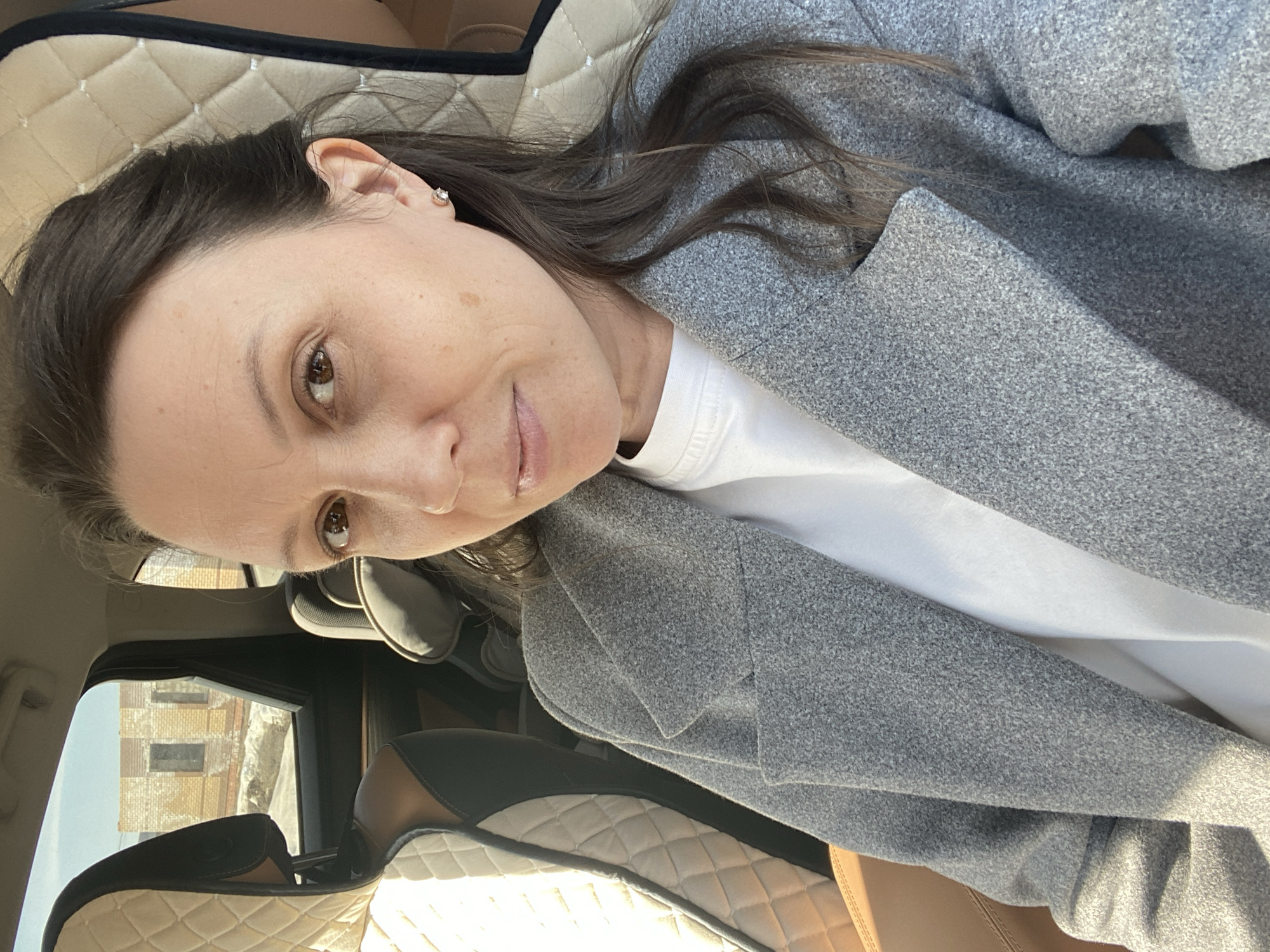 A woman is seated in what appears to be a car, based on the car seat and door visible in the background. She is wearing a white shirt and a grey jacket. Outside the car window, a building is visible, suggesting an urban setting. The building appears to be made of brick or stone. The picture was taken during daylight. The location is Chaikovsky, Russia, based on the presence of distinctive building styles and architectural details often found in that region. The image composition is a selfie-style shot.
