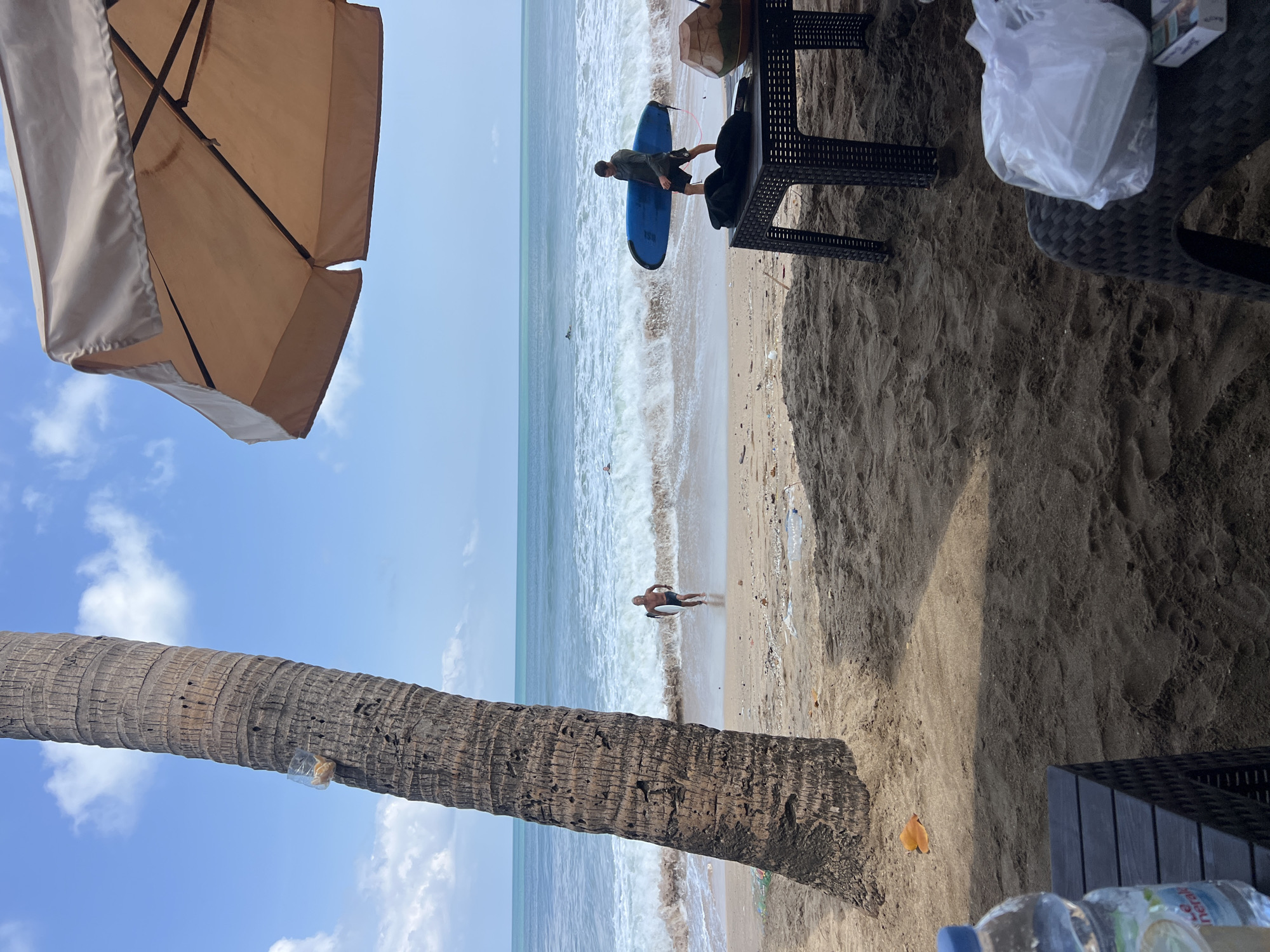 In Kuta, Indonesia, on a sandy beach with a partially obscured palm tree dominating the left foreground, a person is seen wading into the ocean carrying a white surfboard. Further out, a second individual walks along the shoreline holding a blue surfboard. Gentle waves break on the beach under a bright, partly cloudy sky. In the mid-right, a table with a brown umbrella provides shade, and on the table sits a halved coconut and a white bag, possibly containing food. The sand is littered with small pieces of debris.
