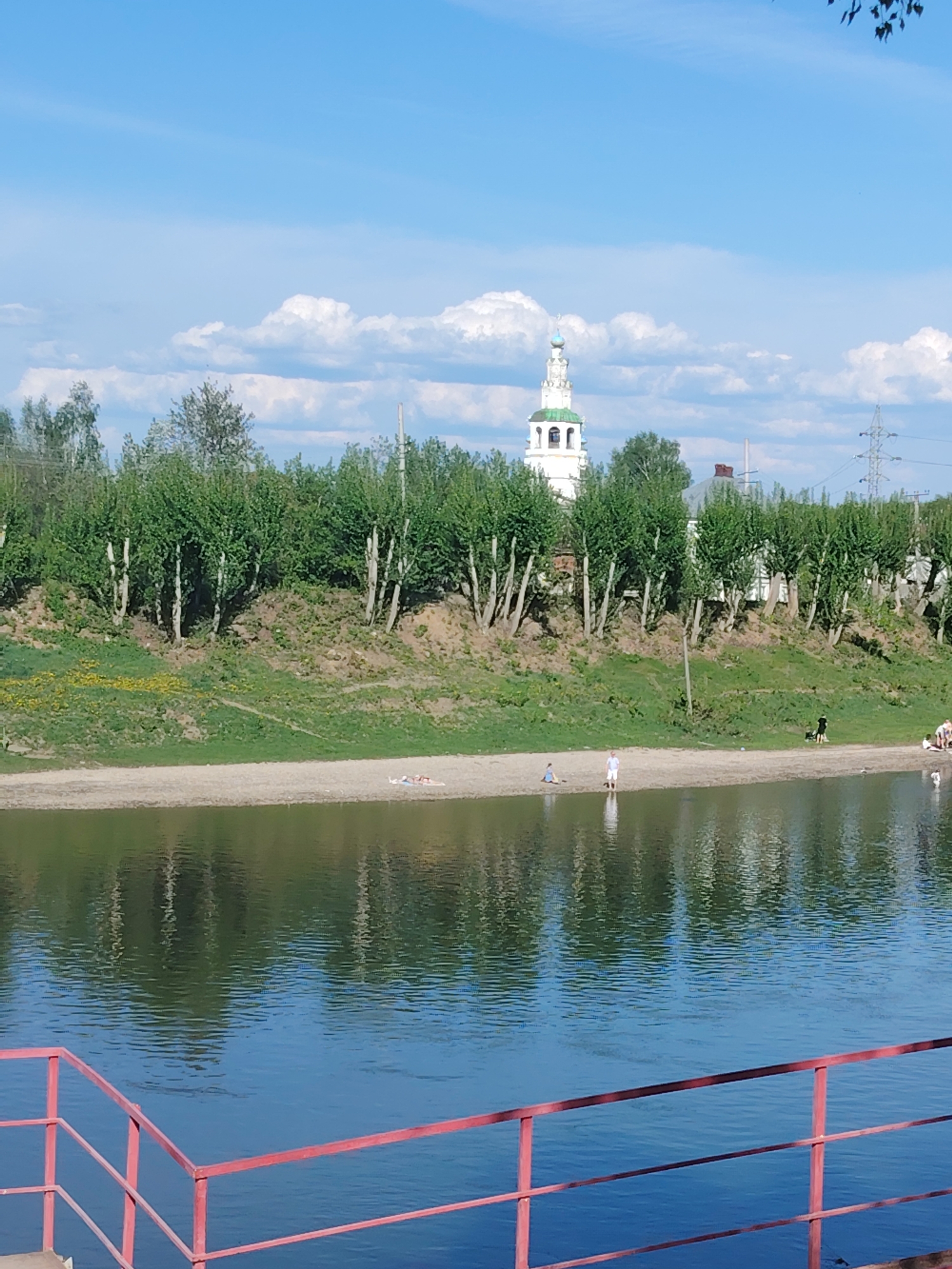 The image depicts a riverside scene in Kungur, Russia. The foreground features a body of water, likely a river, with red railing along the edge. Beyond the water is a sandy bank with scattered individuals, including a person standing in the water. On the raised bank behind the beach, there are trees, grass, and vegetation. In the background, a white building with a tall, ornate spire can be seen amongst the trees, likely a church or religious structure. The sky is blue with puffy white clouds. Power lines are visible on the right side of the background.