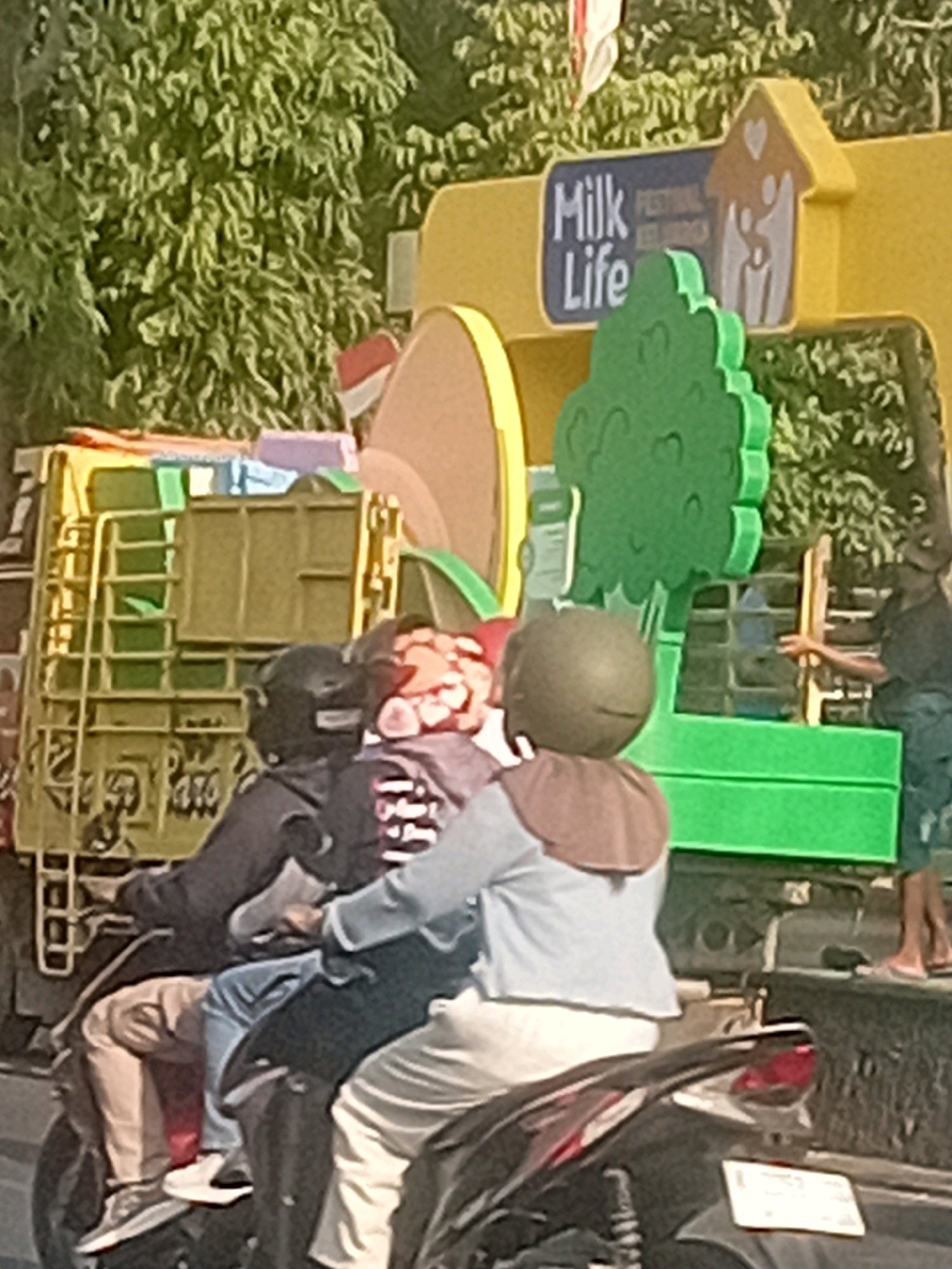 The image depicts a sunny outdoor scene in Kudus, Indonesia, featuring a yellow truck adorned with a "Milk Life Festival" banner. In the foreground, three individuals are riding on a motorcycle. The driver wears a black helmet, while the two passengers, one in the middle and one at the back, also wear helmets and are dressed in casual clothing. They appear to be moving through a street or thoroughfare. In the background, lush green trees frame the scene, suggesting a well-vegetated urban environment. The overall impression is one of a busy, possibly festive, day in the city.