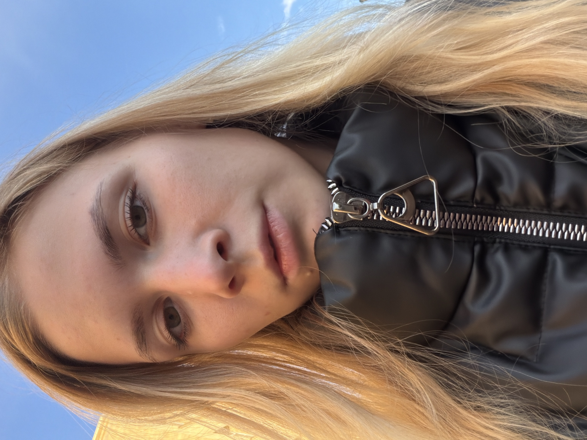 The media file is a close-up, low-angle selfie of a young Caucasian woman with blonde hair. She is wearing a black puffer jacket with a prominent silver zipper. Her face is tilted, and her eyes are looking towards the camera. The background shows a clear blue sky with some hints of clouds. The lighting suggests it is daytime, likely sunny.
The subject's expression is neutral, and the overall mood is casual. The focus is on her facial features and the texture of her hair and jacket. The image appears to be taken outdoors, given the sky in the background. There are no visible text elements in the image. The location context is Izhevsk, Russia.