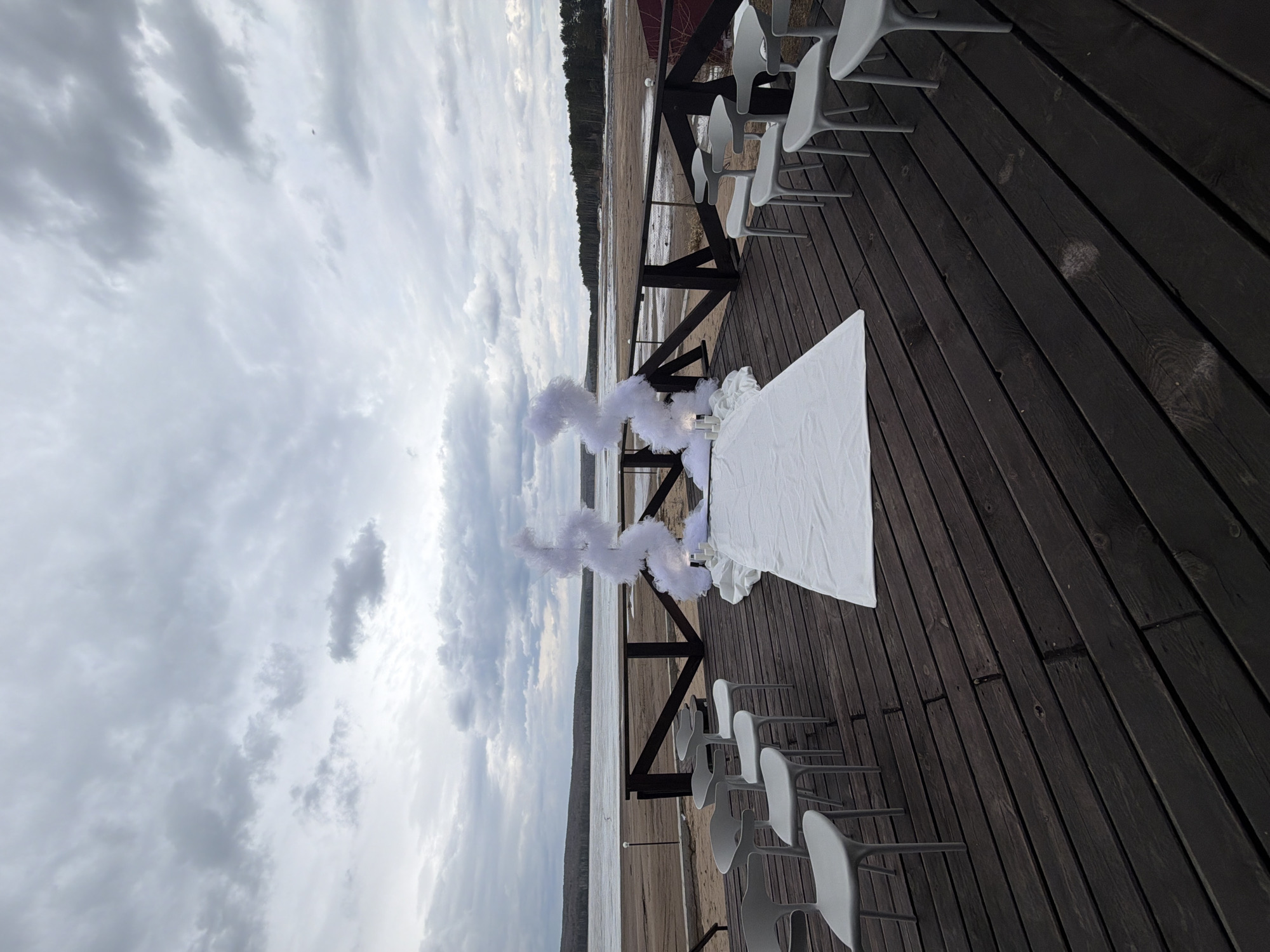 The image depicts an outdoor ceremony setup on a wooden deck, likely prepared for an event such as a wedding. The scene is oriented horizontally, showing a body of water stretching into the distance with a treeline on the far shore.
In the foreground, a dark brown wooden deck serves as the main platform. Several rows of modern, light grey chairs are neatly arranged on the right side of the deck, facing towards a central area. On the left side of the deck, a white-draped table or altar is positioned, adorned with white, cloud-like decorative elements, possibly artificial flowers or fabric arrangements, giving a soft and ethereal appearance. These white decorations are also present further along the railing, creating a sense of an aisle or focal point.
Beyond the deck and its railing, a calm body of water extends, meeting a distant, forested landmass under an overcast sky. The sky is filled with varying shades of grey clouds, suggesting a cloudy day, possibly indicating a muted light or cool weather. There are no people visible in the frame, emphasizing the preparedness and stillness of the setup before an event. The overall atmosphere appears serene and anticipatory. No visible text is present in the image. The setting suggests a picturesque outdoor venue in Chaikovsky, Russia.