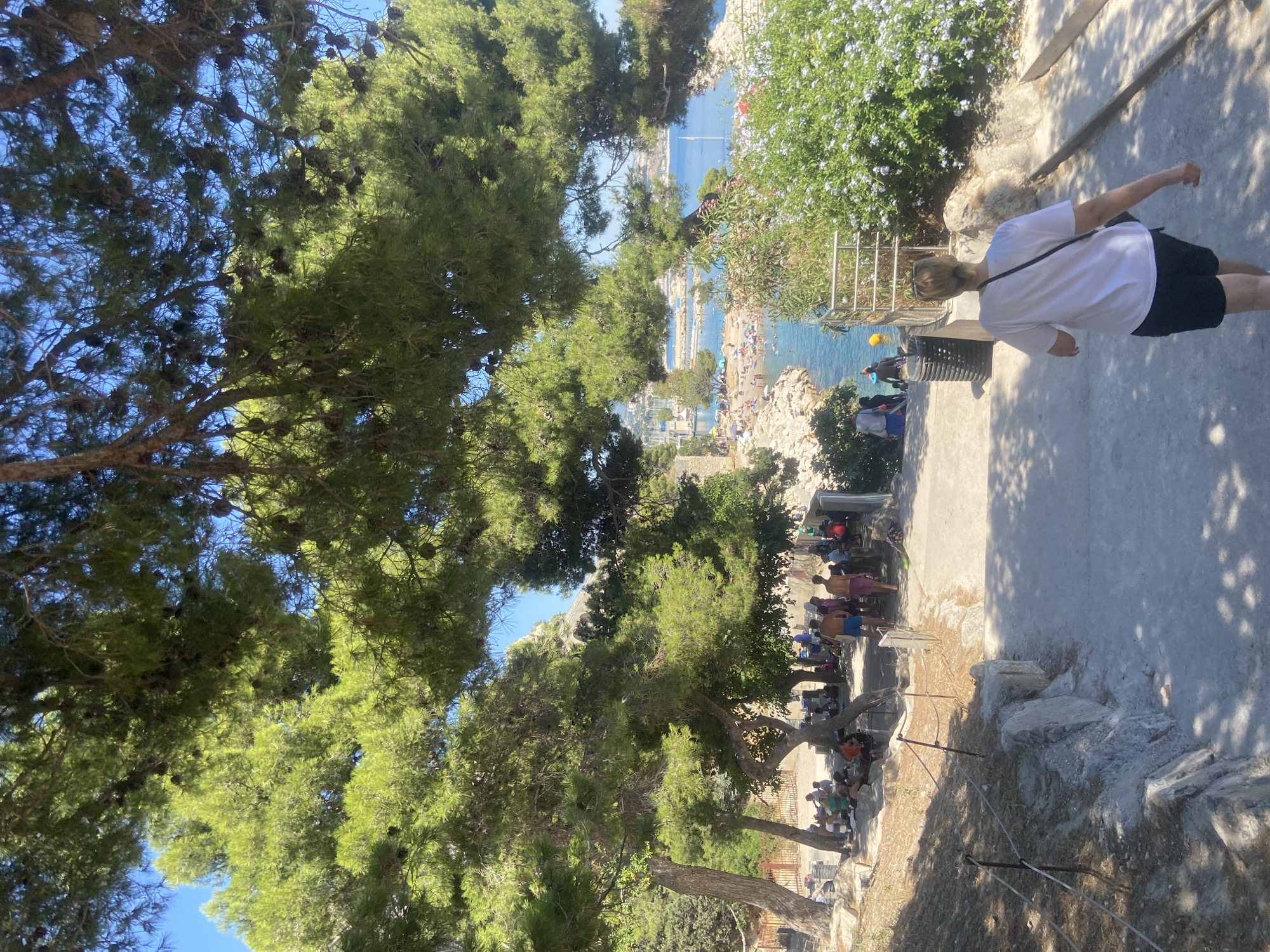 The image shows a view of a crowded beach and bay from a pathway shaded by trees. In the foreground, a woman with blonde hair pulled back is walking down a paved path away from the camera. She is wearing a white t-shirt and dark shorts, with a dark crossbody bag. The path is lined with some rocks and vegetation, and a rope barrier is visible on the left side.
Further down the path, other people are seen walking towards the beach area. The beach itself is full of people sunbathing and swimming in the clear blue water. Boats are anchored in the bay, and buildings are visible on the distant shoreline. Large trees with dense green foliage dominate the upper portion of the image, casting shadows on the path and ground. The sky is a bright, clear blue. The scene appears to be a public recreational area on a sunny day.