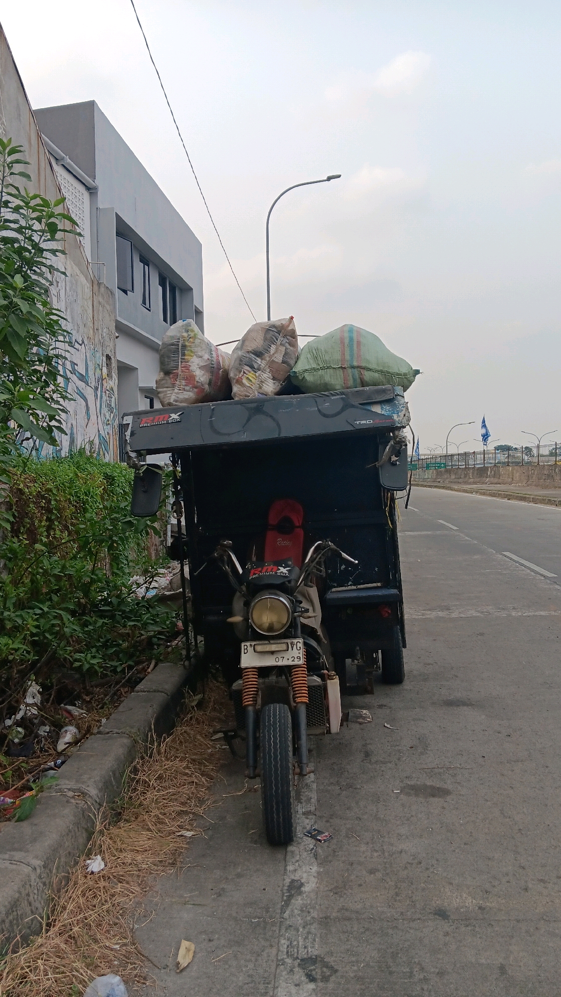 A three-wheeled motorized vehicle, likely a type of "bajaj" or auto-rickshaw, is parked on the side of a road, its bed filled with large, tightly packed bags of what appears to be recyclables or waste. The vehicle has a black canopy over the cargo area, and the front of the vehicle features a motorcycle-like setup with a single headlight and handlebars. A red seat is visible inside the canopy. The license plate, partially obscured, displays "B 796" and "07.29".
The vehicle is positioned on a paved road, with a concrete curb and overgrown dry grass separating it from a patch of green vegetation and a partially visible modern building with grey cladding and dark windows. Graffiti can be seen on the exterior wall of the building on the left. In the background, a street light arcs over the road, and further in the distance, a highway overpass with railings and flags is visible. The sky is overcast. The scene appears to be located in an urban or semi-urban area of Depok, Indonesia, possibly a collection point or a road where such vehicles operate for waste management.