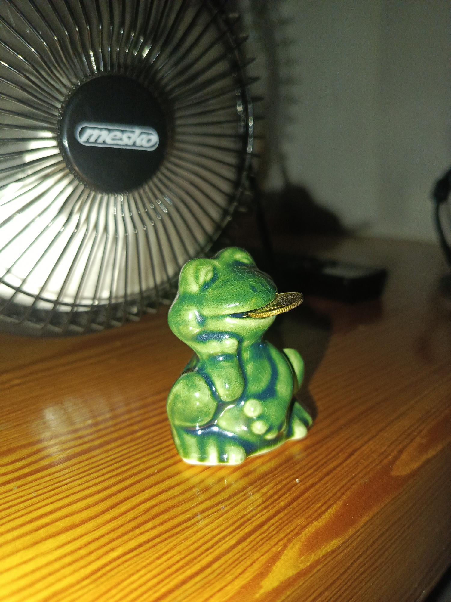 The image shows a small, green, ceramic frog figurine on a wooden surface. The frog is sitting and has a gold-colored coin partially inside its mouth. Behind the frog, a black and silver fan is visible. The fan has a circular grill and a central black dome with a white logo that reads "mesko". The wooden surface appears to be a tabletop with a visible wood grain pattern. The lighting in the image is dim, with a spotlight effect on the fan and the figurine. The background is out of focus and appears to be a wall. The scene is likely indoors, possibly in a home or office. There is no indication of the specific location in Bolesławiec, Poland, as this is not an identifiable landmark.