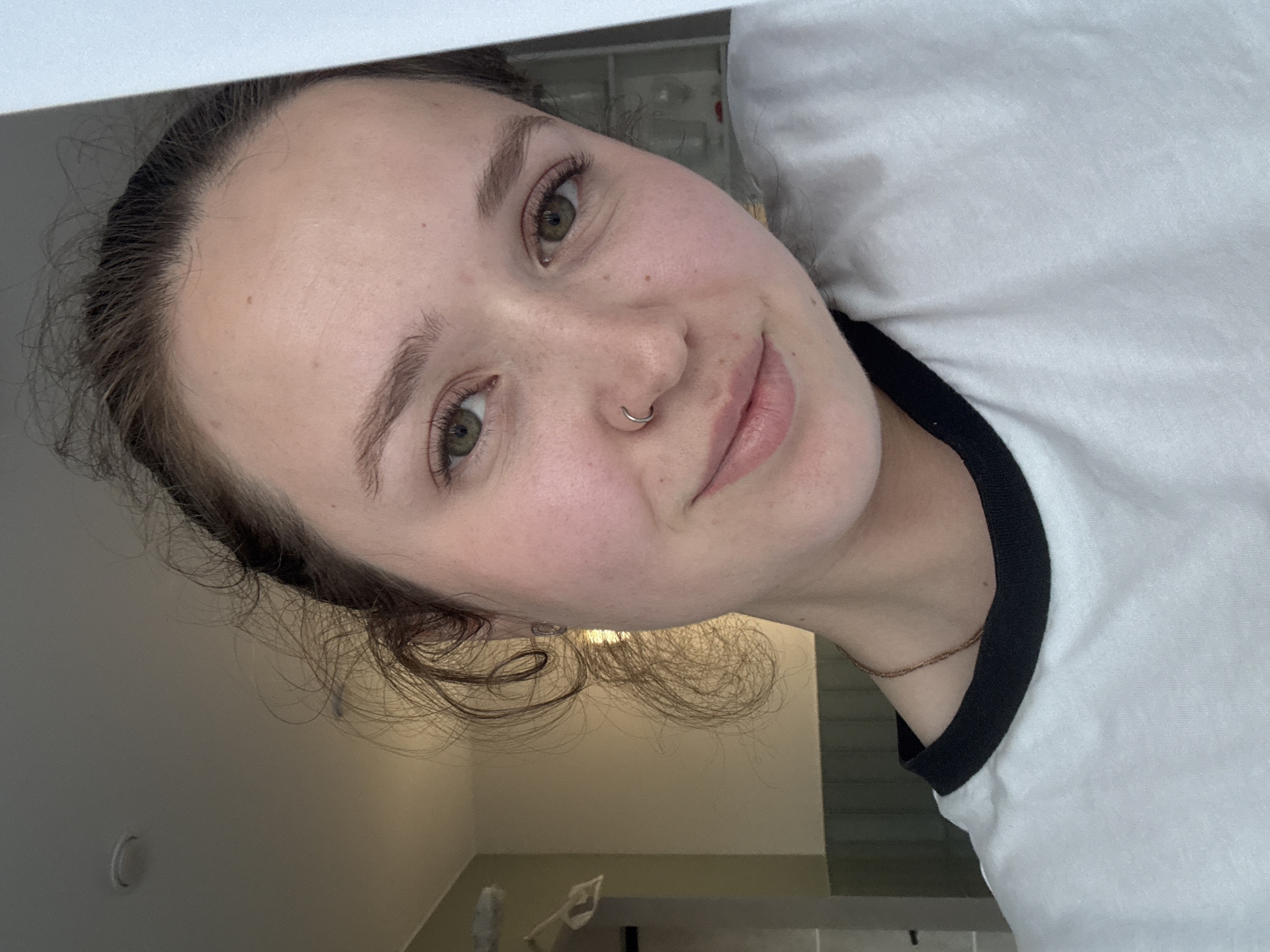 A close-up selfie of a young woman with fair skin, light brown hair pulled back from her face, and green eyes. She has a nose piercing and is wearing a white T-shirt with a black collar. The background is blurry and indistinct, showing hints of a wall, lighting fixture, and possibly a bathroom or similar interior space. It is not possible to determine the location in the city of Izhevsk, Russia.