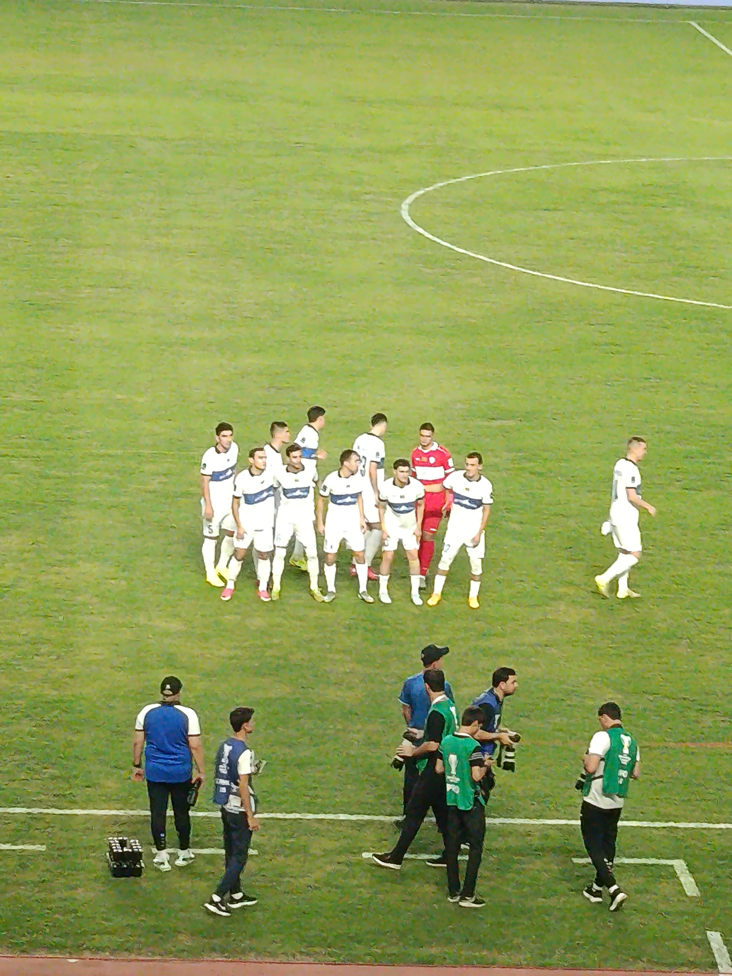 A group of men in soccer uniforms stand on a vibrant green grass field. A semicircle is marked on the field with a white line. In the foreground, several men in dark clothing and vests, some carrying cameras, are present. The field appears to be part of a sports stadium, possibly within the city of Arkadag, Turkmenistan, given the context. The overall lighting suggests it is daytime.