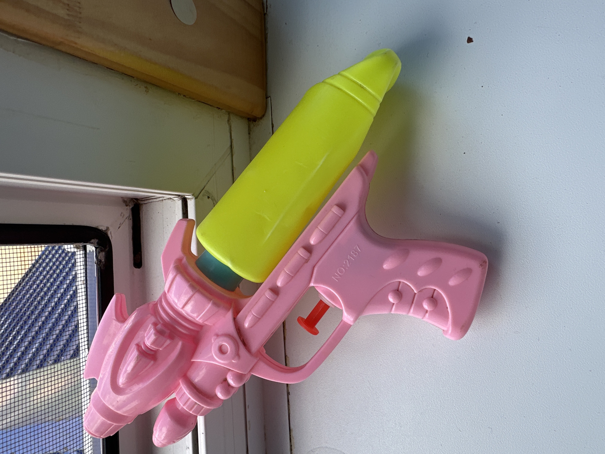 A pink plastic toy water gun is positioned on a white surface. The water gun has a rocket-shaped body and a yellow reservoir. The image is cropped to show the water gun and a hint of a window with a screen in the background. There is no indication of the city of Мариинский, Russia, or any specific urban environment.