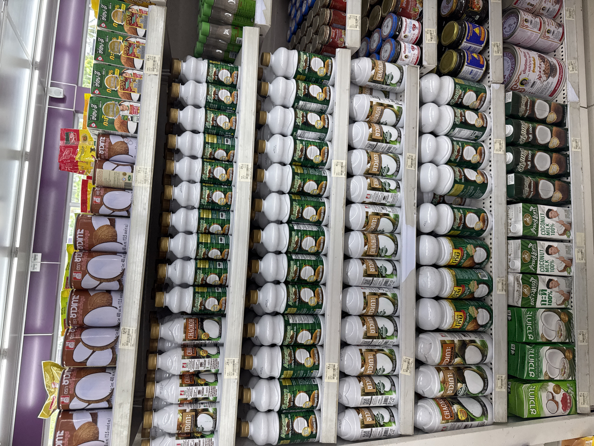 The image shows shelves stocked with various brands of coconut milk and related products in what appears to be a grocery store or market setting. The shelves are organized vertically, with different brands and sizes of coconut milk lined up. The products include coconut milk in cartons and bottles with recognizable brands like Ampawa, Chao Koh, and Luicla. There are also boxes of a drink called "Oreo Dee," and some small jars, possibly sauces or condiments. Price tags are visible on the shelves. The bottles are packaged with white caps. The location is likely a shop in Thong Sala, Thailand.