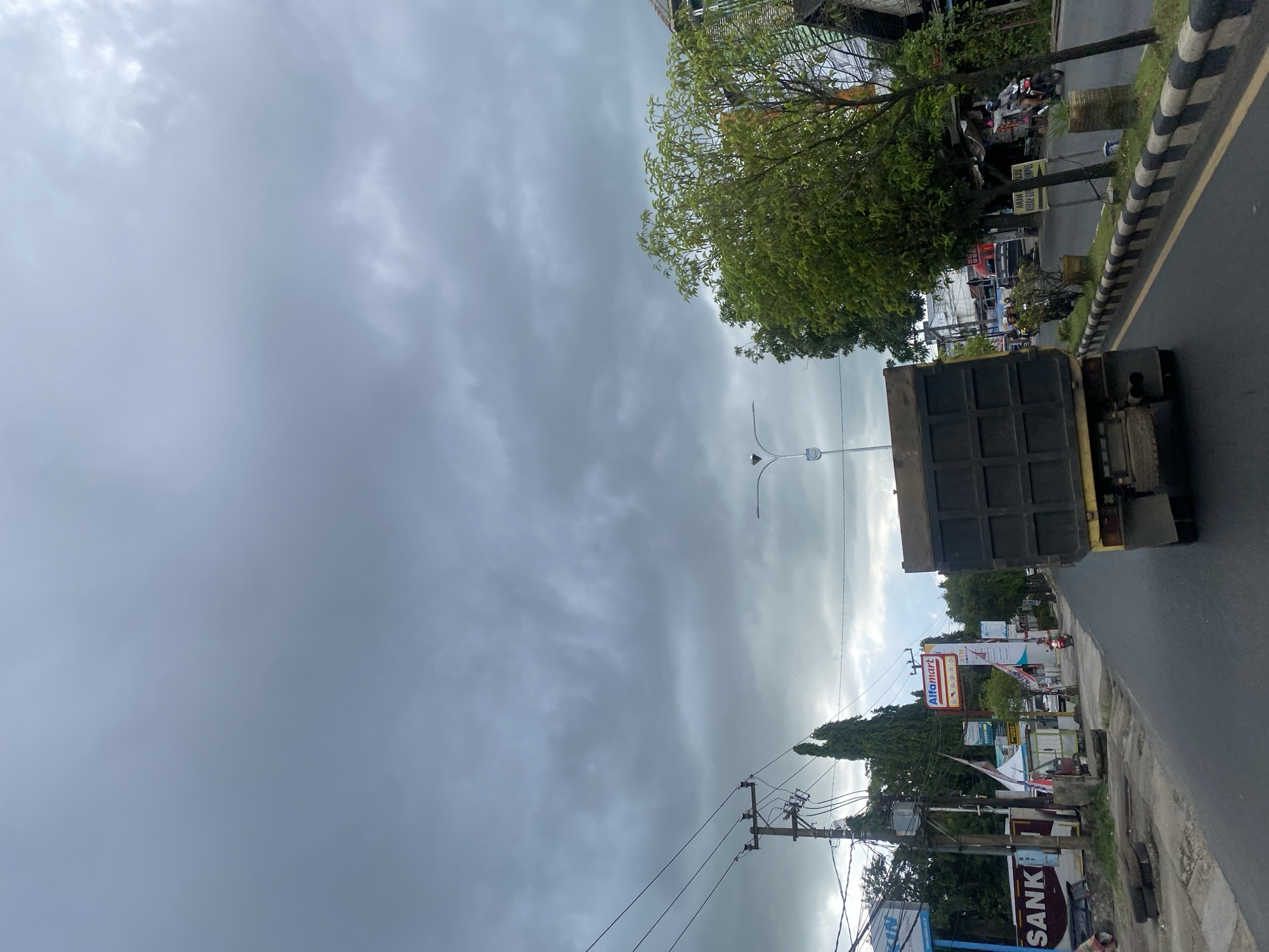 The image depicts a street scene in Sungai Paring, Indonesia, under a cloudy, overcast sky. A yellow dump truck with a black bed is the primary subject, driving away from the camera on the right side of a paved road. The truck has dual exhaust pipes. The road has a distinct black and white striped curb. To the left of the road, a sidewalk area is visible, with stacked tires and various structures including a utility pole with wires. Signs are present, including one for "Alfamart" and another with the text "SANKI". There are trees along the roadside, with one prominent green tree on the right side of the image. A few motorcycles and a three-wheeled vehicle are also visible, suggesting a moderately busy urban or suburban environment. The overall atmosphere is muted due to the cloudy weather.