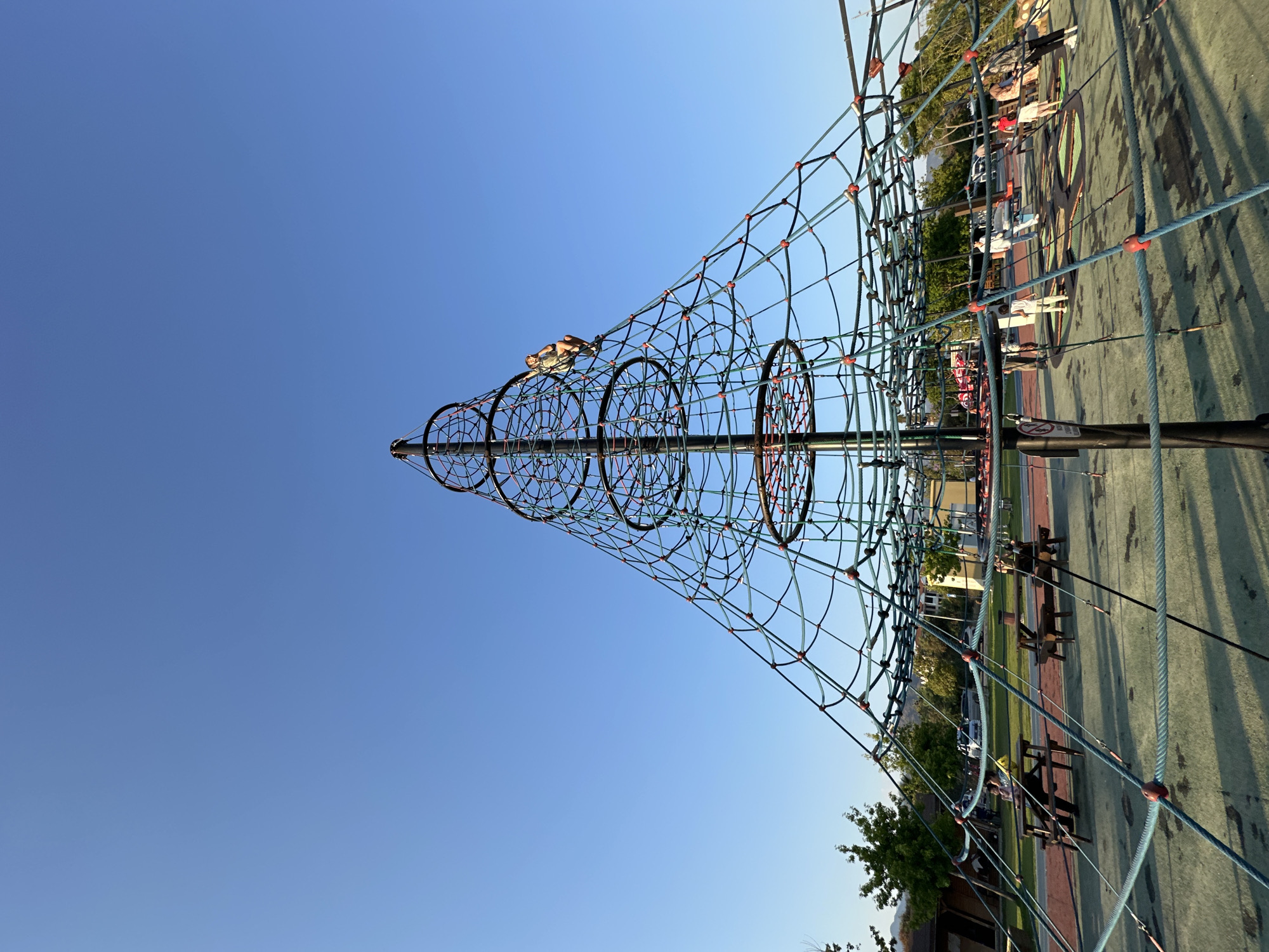 This is a wide-angle, low-angle shot of a large, conical rope climbing structure at a playground, likely in Fethiye, Turkey, given the blue sky and the style of buildings in the background. The structure dominates the frame, with a central dark pole and a net of blue and green ropes forming a pyramid shape. Red connectors are visible at the intersections of the ropes. A person, appearing to be a child, is climbing on the upper section of the net.
In the foreground and mid-ground, picnic tables and a soft, green playground surface are visible. Several other people, including adults and children, are scattered around the playground. Some are walking, others are standing near playground equipment, and a few are seated at the picnic tables. Trees and buildings are visible in the background, suggesting a park or recreational area within a developed setting. The sky is a clear, vibrant blue, indicating good weather. The lighting suggests it is late afternoon or early evening, with the sun casting some shadows.