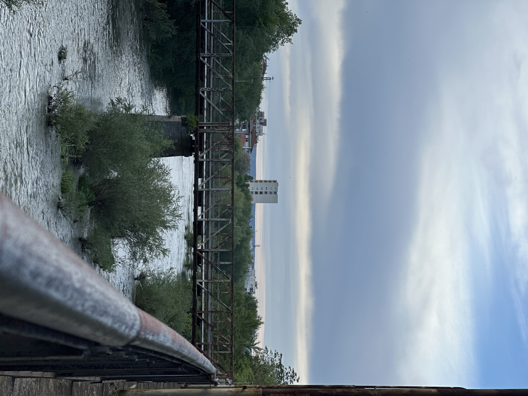 The image depicts a view from a rusty metal bridge in Lovech, Bulgaria, overlooking the Osam River. The river flows with turbulent water around small islands of vegetation. Another similar, parallel bridge can be seen next to the one the photo is taken from. Beyond the immediate riverbank vegetation, the cityscape of Lovech is visible, including residential buildings and a tall, blocky structure. The sky is overcast with banded clouds. The photograph is taken from an elevated perspective, looking out over the bridge's structure and the surrounding scenery.