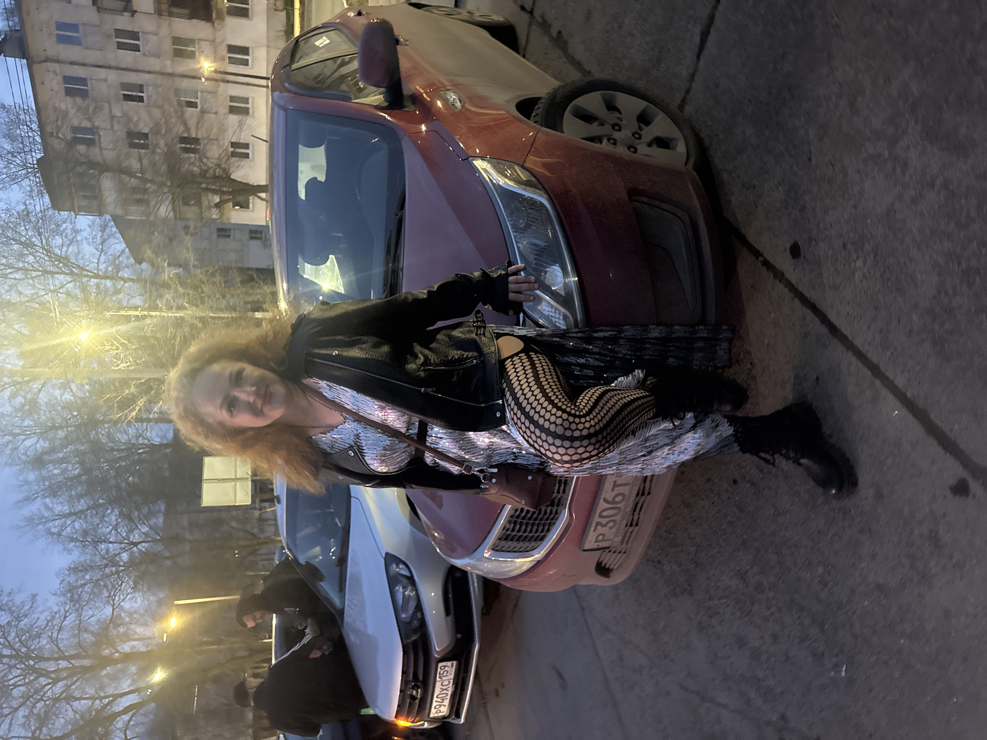 A photograph, oriented vertically, depicts a woman leaning against a maroon car parked on an urban street in Chaikovsky, Russia. The woman has reddish-blonde hair and is wearing a black leather jacket, a silver sequined dress, and black fishnet stockings. She is also carrying a brown shoulder bag.
The car, identified by license plate P306TX, is parked next to a white car with license plate P940XC159. Two people are visible near the white car.
In the background, an apartment building is visible, along with trees and streetlights. The light suggests it is either dusk or dawn. The image is captured from ground level. The location is in Chaikovsky, Russia.