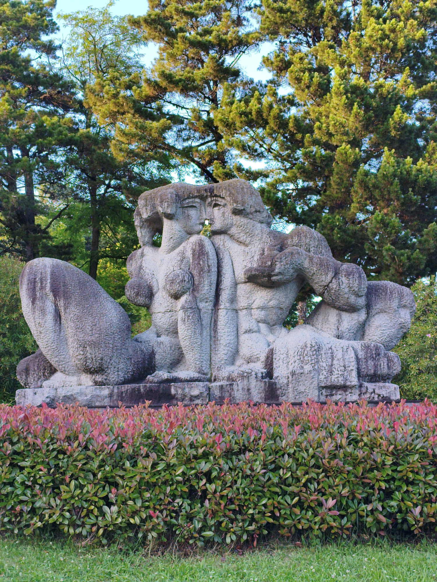 The image shows a large, abstract stone sculpture of multiple human figures in a park setting. The sculpture is made of rough-textured gray stone and depicts intertwined figures with simplified features. In front of the sculpture, there is a neatly trimmed hedge with red and green foliage. Behind the sculpture, tall trees with green leaves are visible against a clear blue sky. The ground in the foreground is covered with green grass. The sculpture is located in the Arboretum botanical garden in Sochi, Russia. There are no people visible in the image.