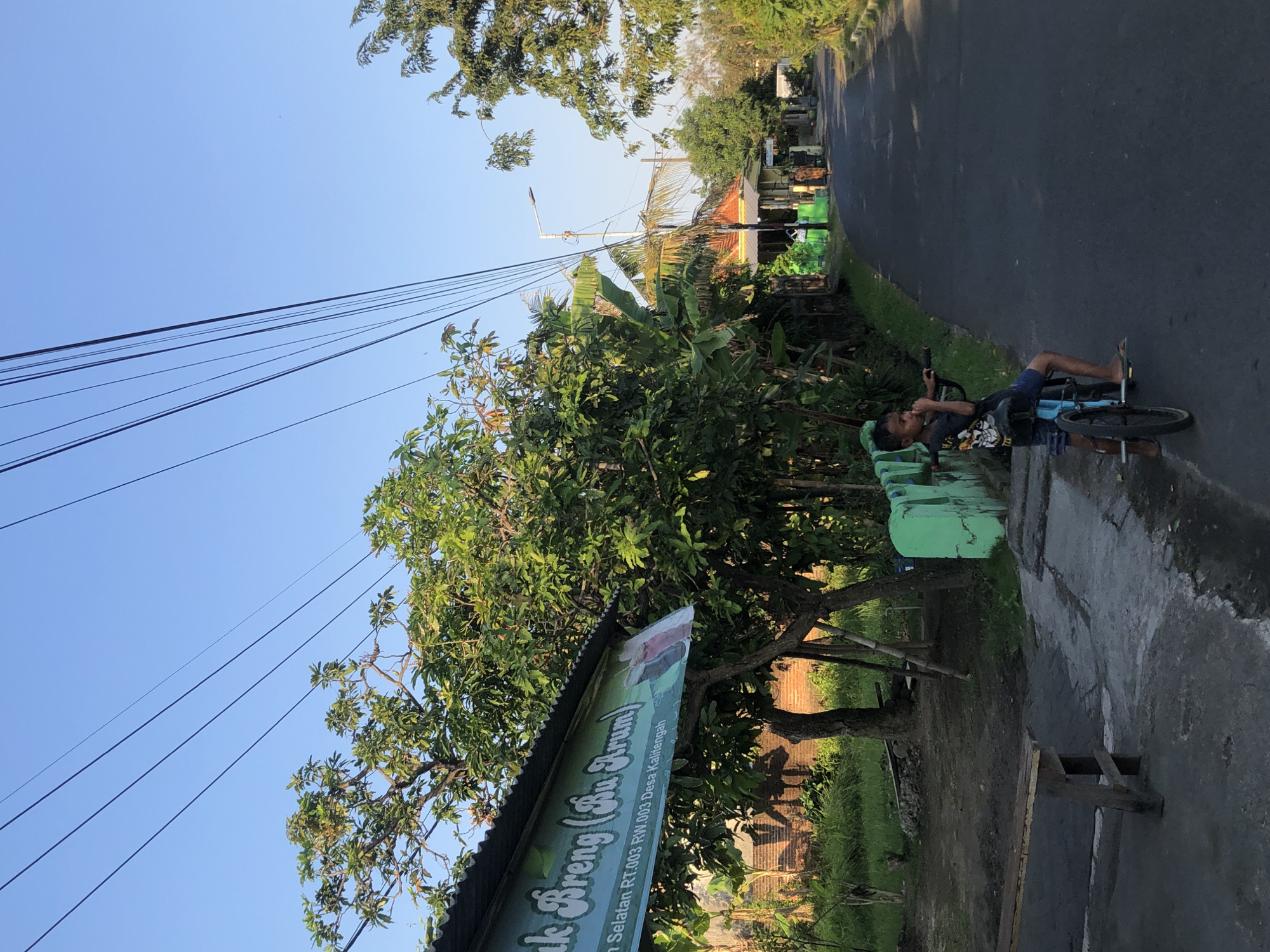 In Kalitengah, Indonesia, a young boy is seen standing on a road with his bicycle. The boy is wearing a dark t-shirt and shorts. He is looking towards the right side of the frame. The bicycle is blue and has a training wheel. In the background, there are trees, houses, and a street lamp. The sky is clear and blue. A sign that reads "ak Breng (Bu Arum)" is visible on the left side of the image, above a wooden bench. The road appears to be an asphalt surface. A person is walking in the distance on the right side of the road. A green wall with decorative elements is present on the left side of the road.