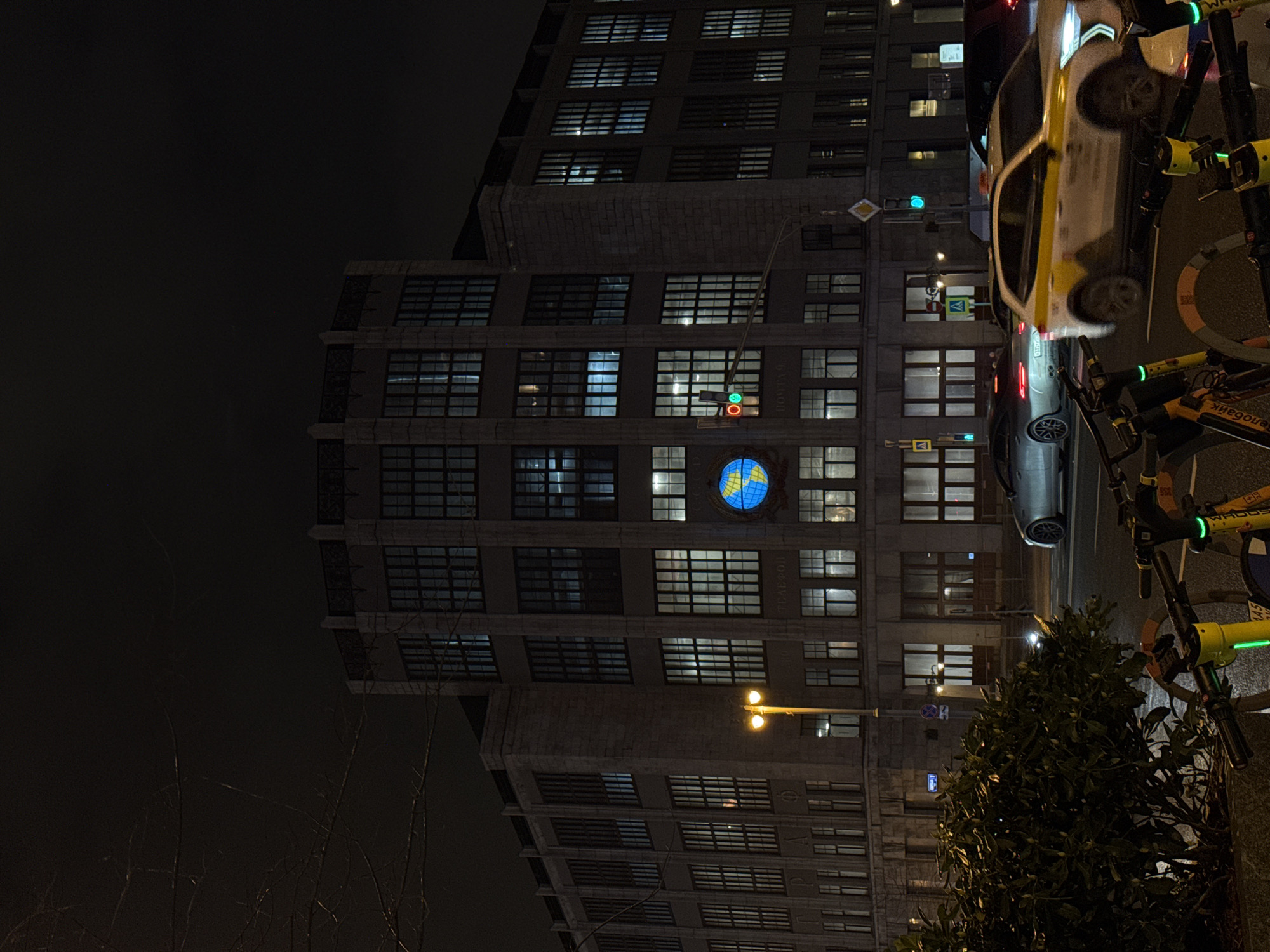At night, an eye-level, medium shot depicts the Central Telegraph Building in Moscow, Russia. The building is tall and boxy, with rows of windows. A lit globe is visible on one of the floors. Street lights illuminate the front of the building. A street with cars and scooters is to the right. A small tree is also present on the right side of the image. The sky is dark.