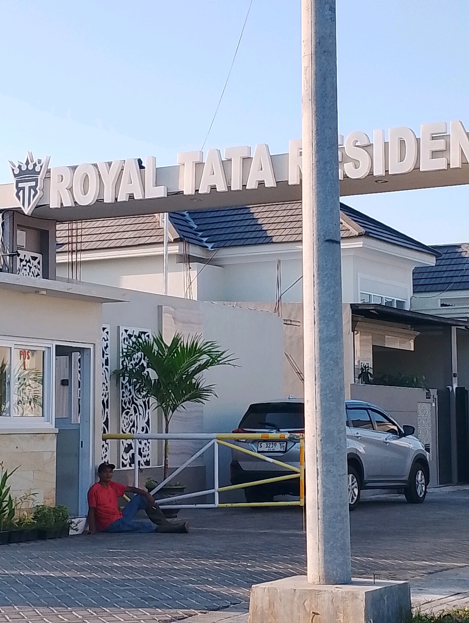 The image shows the entrance to a residential complex named "ROYAL TATA RESIDENT" in Probolinggo, Indonesia. A man in a red shirt and blue jeans is sitting on the ground near the entrance gate. A silver SUV with license plate S 1219 YG is parked behind the gate. The entrance is marked by a large white archway with the residential complex's name in prominent white lettering. The area appears to be a paved driveway with decorative patterned walls and a small palm tree. The sky is clear and blue, suggesting it is daytime.