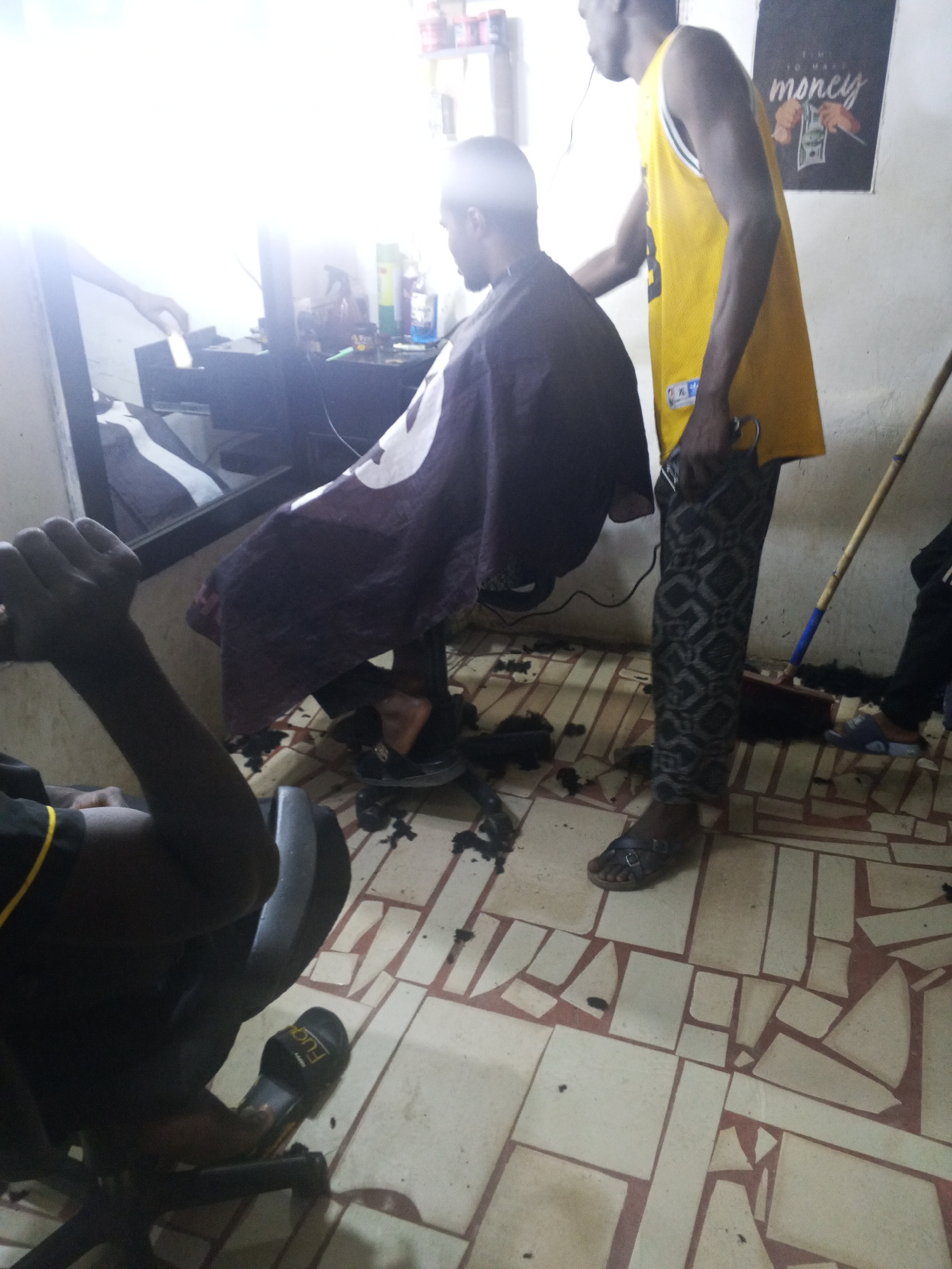 The image shows the interior of a barbershop in Gwong, Nigeria. A barber in a yellow basketball jersey and patterned pants stands beside a seated customer draped in a maroon-colored cape. The barber holds a pair of scissors in his hand. Hair clippings are scattered on the tiled floor. A second individual is partially visible in the foreground. The environment appears utilitarian and modestly furnished. A mirror and shelves with products are positioned behind the customer. A poster with the word "money" is visible on the wall.
