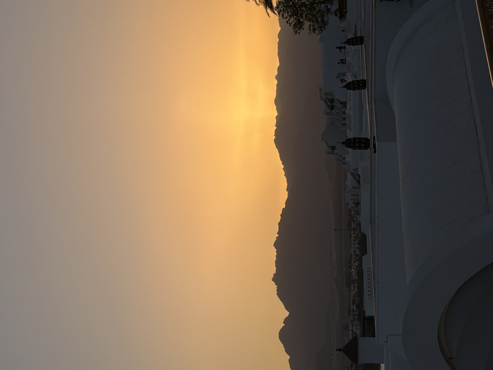 The image, when rotated clockwise to its correct landscape orientation, captures a stunning sunset or sunrise over Sharm El Sheikh, Egypt. The sky dominates the left and central parts of the frame, showcasing a vibrant gradient of warm colors. A brilliant golden-yellow light radiates from just above the horizon, suggesting the sun's position, fading into softer oranges and muted greyish-orange hues towards the left and top.
A dark, rugged mountain range stretches across the middle ground, forming a prominent silhouette against the luminous sky. Its jagged peaks and contours are distinctly outlined by the backlighting. Below the mountain range, in the far distance, faint specks of light are visible, indicating a distant settlement or urban area.
In the foreground and extending along the right side of the image, distinctive white architectural structures are visible. These buildings feature smooth, curvilinear forms, arches, and railings, consistent with resort or hotel architecture. Several decorative, dark lanterns hang from these structures, which are partially cast in shadow due to the angle of the sun. A dark silhouette of foliage, possibly palm fronds, can be seen in the upper right corner.
The scene conveys a sense of peacefulness and natural beauty, characteristic of a desert landscape at either dawn or dusk. There is no discernible activity taking place, and no visible text in the image.