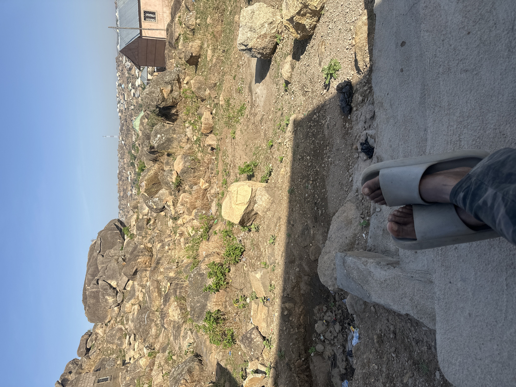 The image, taken outdoors in Jos, Nigeria, shows a view from a high vantage point overlooking a densely populated urban area. The foreground features a concrete structure with a person's legs and feet visible, shod in light grey slip-on sandals. The environment consists of rocky terrain with sparse vegetation and scattered debris. A building with a light-colored exterior and corrugated metal roof is positioned near the top of the frame. The urban landscape in the background is characterized by a multitude of closely packed buildings. The sky is clear and blue.