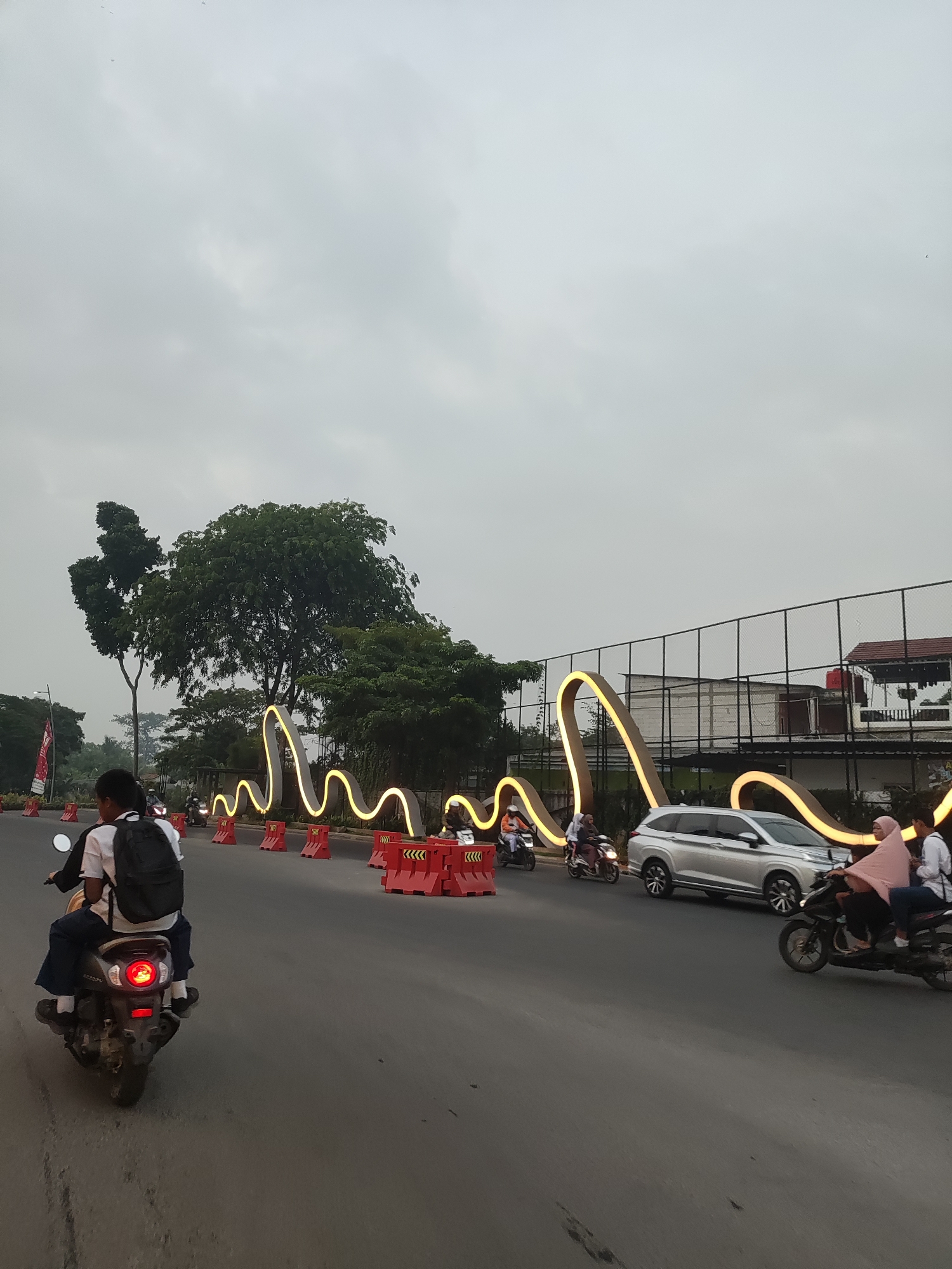 The image shows a street scene in Telagaasih, Indonesia, on a cloudy day. In the foreground, a person rides a motorcycle with a backpack, wearing a white shirt and dark pants. Behind them, several other motorcycles are visible, some with passengers. A silver minivan is driving on the road. Along the side of the road, there are bright, wavy light installations with red traffic barriers. In the background, there are trees and a fenced area with buildings. The overall environment appears to be an urban or suburban street.