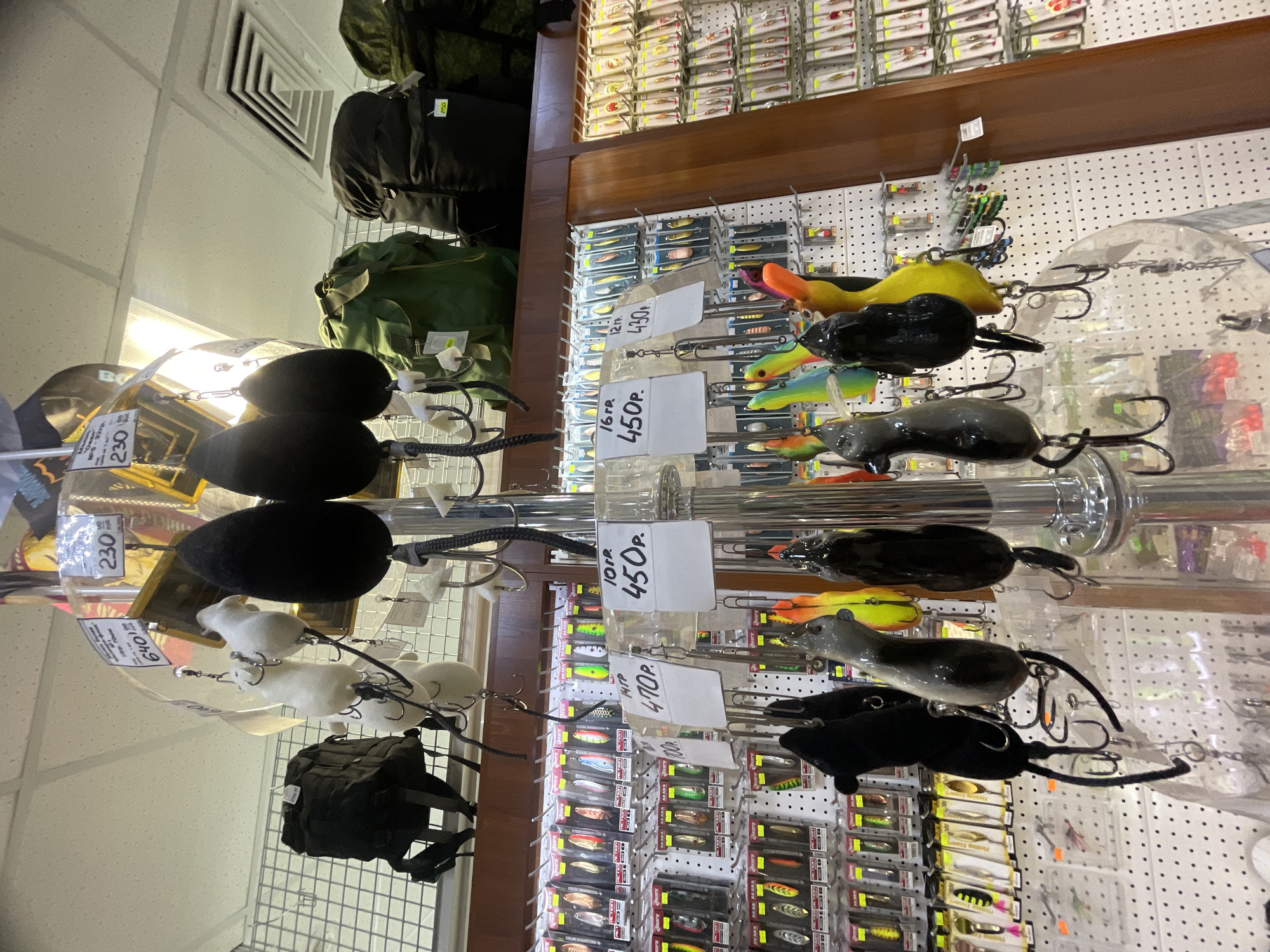 Here's a detailed description of the image:
**General Scene:**
The image appears to be taken inside a store that sells fishing lures and possibly other outdoor or tactical gear. The angle is unusual, taken looking upwards at merchandise displayed on shelves and hanging racks.
**Objects:**
* **Fishing Lures:** The main focus is on a display of fishing lures. These lures are designed to resemble mice or other small animals. They come in various colors: black, gray, white, yellow/orange. The lures are hanging on a rack.
* **Packaging:** Rows of packaged fishing lures are visible in the background, neatly arranged on shelves.
* **Price Tags:** Small white price tags are attached to the lures on the hanging rack. The price tags display the prices in Russian currency (Rubles). The prices visible include 230, 430, 450, and 470. Also on the price tags is information like the weight of the lures, in grams: 5-32гр, 12 гр, 10гр, 16гр.
* **Gear/Bags:** Black and green backpacks or bags are hanging above the lure display.
* **Racks:** The lures are displayed on a metal rack, with a central post and arms extending outwards.
**Environment:**
* **Retail Store:** The setting is clearly a retail environment.
* **Lighting:** The lighting is bright, possibly from overhead fluorescent fixtures.
* **Walls:** The walls are light-colored and appear to be made of panels.
**Location Inference:**
Based on the Cyrillic text and the Ruble pricing, the location is most likely in Russia. Given the OCR text, it can be inferred that the fishing lures are sold in Krasnoyarsk, Russia.