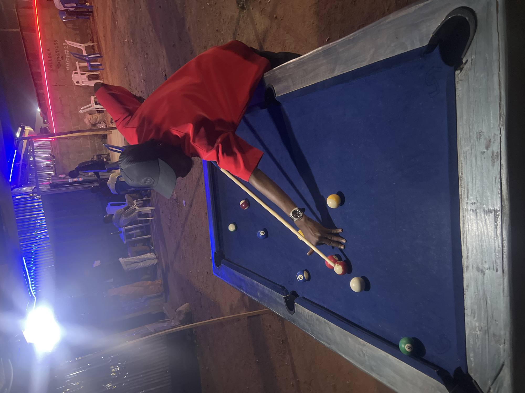 A man in a red shirt and grey cap is playing pool on a blue felt pool table, located in what appears to be an outdoor entertainment space in Gura Top, Nigeria. The pool table has several balls on it, and the man is holding a pool cue with his right hand while positioning his left hand on the table to steady his aim. There are people sitting at tables and chairs in the background, under a structure illuminated with blue and red neon lights. The ground around the area appears to be dirt.