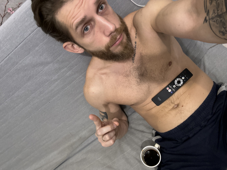 An adult male is depicted in an indoor setting, lying supine on a light gray, ribbed or striped bedsheet. The precise city location of Saint Petersburg, Russia, cannot be determined from the visual content of the image.
The male subject possesses light skin, blue eyes, a full brown beard, and dark hair styled, with some pulled back, visible in the upper right. He is shirtless, exposing his torso, and wears dark blue athletic or pajama shorts. A simple gold-colored band is present on the ring finger of his left hand. Tattoos are visible on his body: a partially obscured circular design on his left bicep/shoulder, and Cyrillic text across his upper chest, beginning "Обжоры с..." (translating to "Gluttons from...").
Objects include a white ceramic mug, containing a dark liquid, likely coffee, positioned on the bedding to the left of his abdomen. A black television remote control, bearing the "HONOR" brand logo and buttons for streaming services such as Netflix and YouTube, rests horizontally on his abdomen.
The male subject's left hand is raised, with his index finger extended, pointing towards the upper left. He is looking directly at the camera, implying a self-portrait (selfie). His right arm is bent, with his hand out of frame, consistent with holding a camera.
The environment is consistent with a bedroom. A small portion of a decorative element, possibly a headboard or wall art with a floral pattern, is visible in the upper right background. A white cable or cord is also partially visible near the top right.