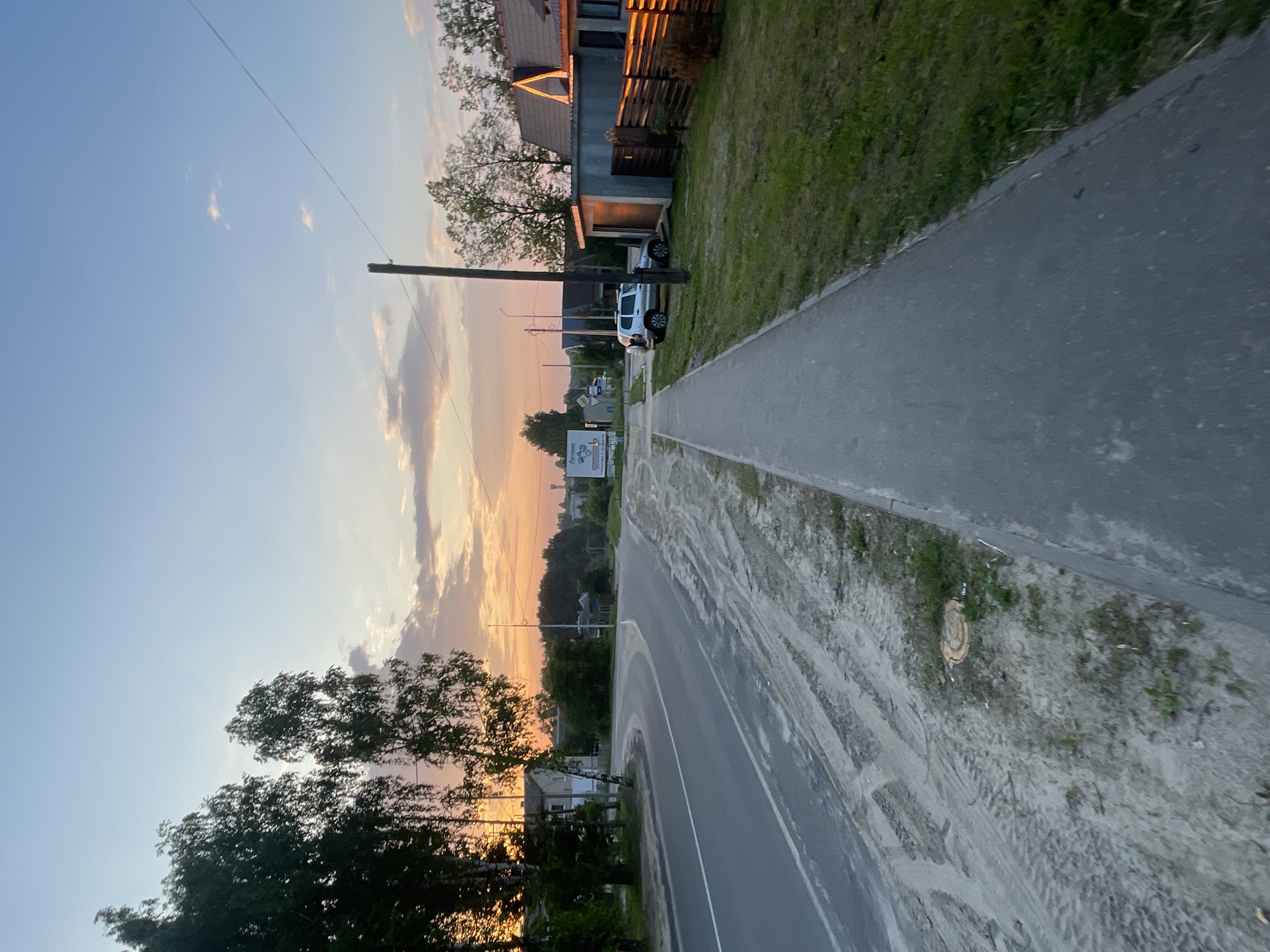 The image depicts a paved sidewalk running alongside a road with a sandy, tire-tracked shoulder. The scene is set in a rural or suburban area, possibly Красные Баки, Russia, given the context. The time of day is sunset, evidenced by the warm, orange and pink hues in the sky and the long shadows cast.
On the right side of the sidewalk, there is a well-maintained lawn bordering a wooden fence. Beyond the fence, a house with an orange-tiled roof is visible, catching the last rays of the sun. A small, attached garage is next to the house, and a silver SUV is parked in front of it. A tall wooden utility pole stands near the sidewalk, with a power line extending across the frame.
Across the road, on the left side, is a paved road with a white lane marking. The shoulder of the road is composed of sand and dirt, with visible tire tracks. Several trees with dark green foliage are present, and a building, possibly a house, is situated behind them. Further in the background, more trees and some structures suggest a small settlement. A billboard with text and graphics is visible in the distance on the left. The overall environment appears quiet and tranquil, characteristic of a rural sunset.