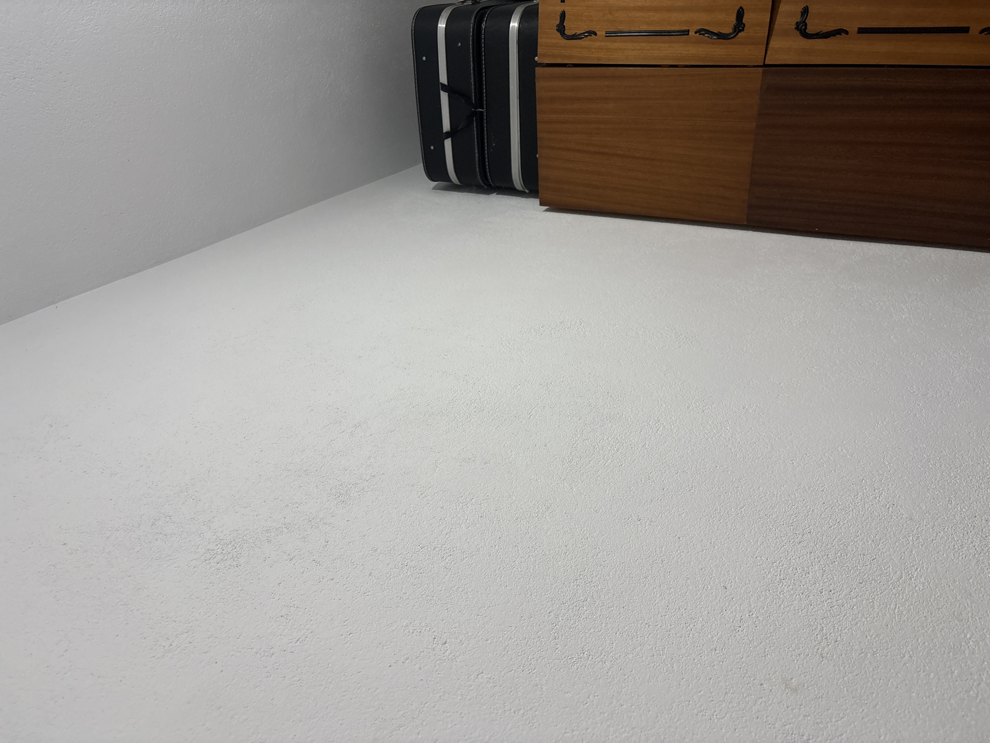 The image shows a corner of a room with a white textured floor and walls. Along the wall on the right side is a piece of wooden furniture, likely a dresser, with visible drawers and metal handles. Adjacent to the dresser, closer to the corner, stands a black suitcase with silver accents. The location is not possible to determine from the image.
