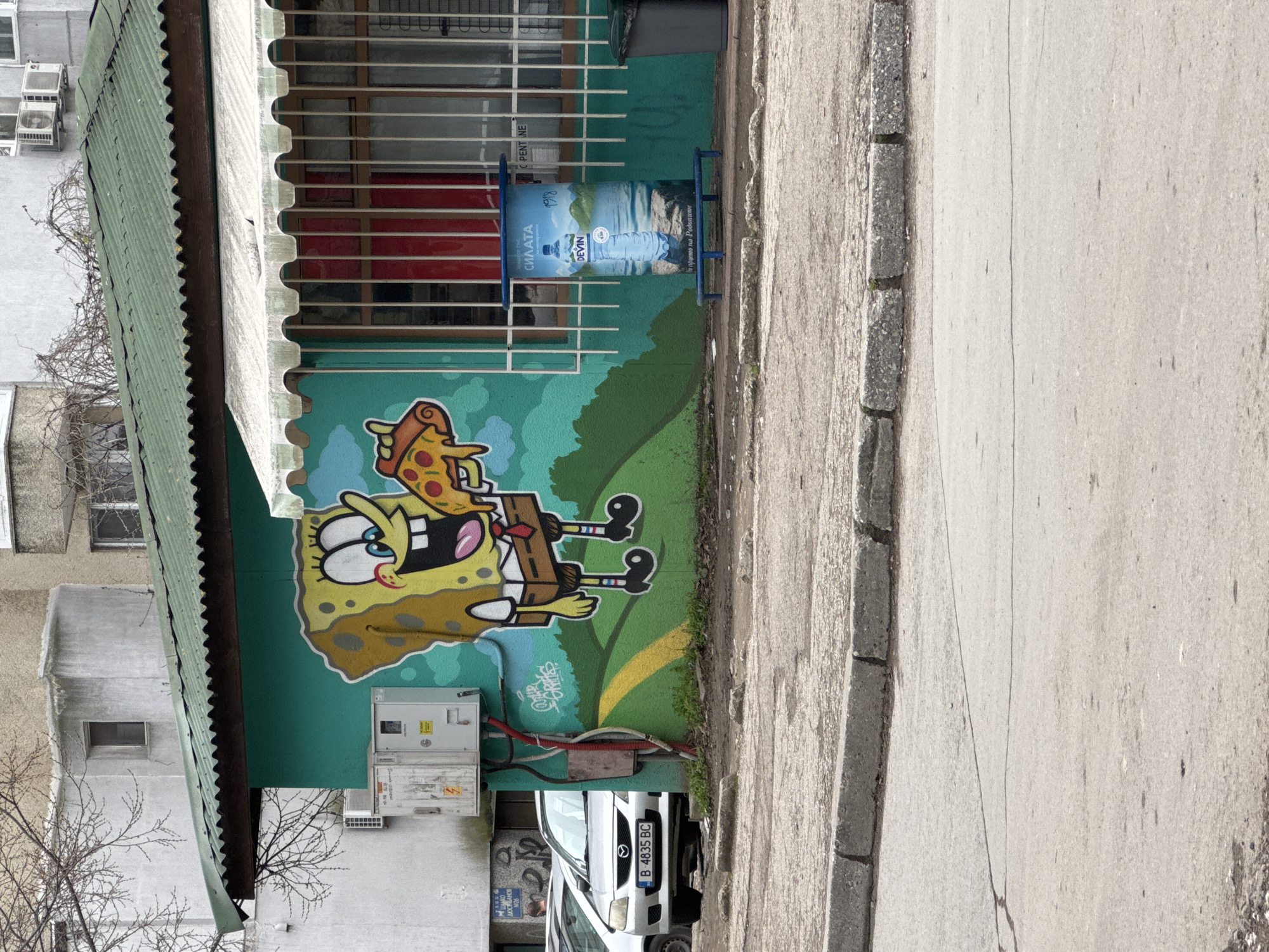 The image shows a street view in Varna, Bulgaria, featuring a mural of Spongebob Squarepants eating pizza on a turquoise wall. The mural is on the side of a building with a green awning and a security grate. A "Devin" water advertisement is visible next to the mural. Below the mural are electrical boxes and a white Mazda car with license plate "B 4835 BC" parked on the side of the street. The sidewalk is concrete, and bare tree branches are visible in the background. The scene appears to be daytime.