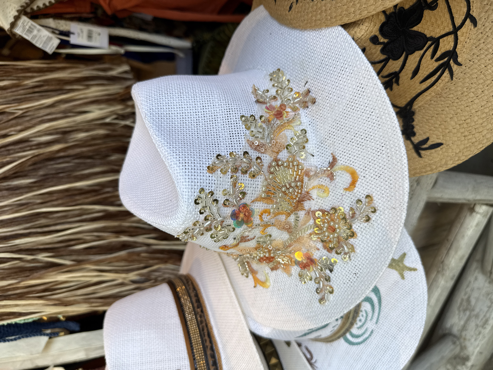 This image shows a close-up of several hats for sale at a street market in Thassos, Greece. The foreground is dominated by a white straw hat with intricate beadwork and sequin embellishments in shades of gold, orange, and pink, arranged in a floral pattern around the crown. To the left and slightly behind, another white straw hat is visible, featuring a decorative band with woven brown and gold elements. Beneath these, parts of two other straw hats are visible; one is tan with black floral embroidery, and another appears to have a green circular design. The background consists of a display of natural fibers, possibly dried grasses or reeds, and some hanging clothing items with tags. The lighting suggests an outdoor setting during the daytime, likely in a tourist area.