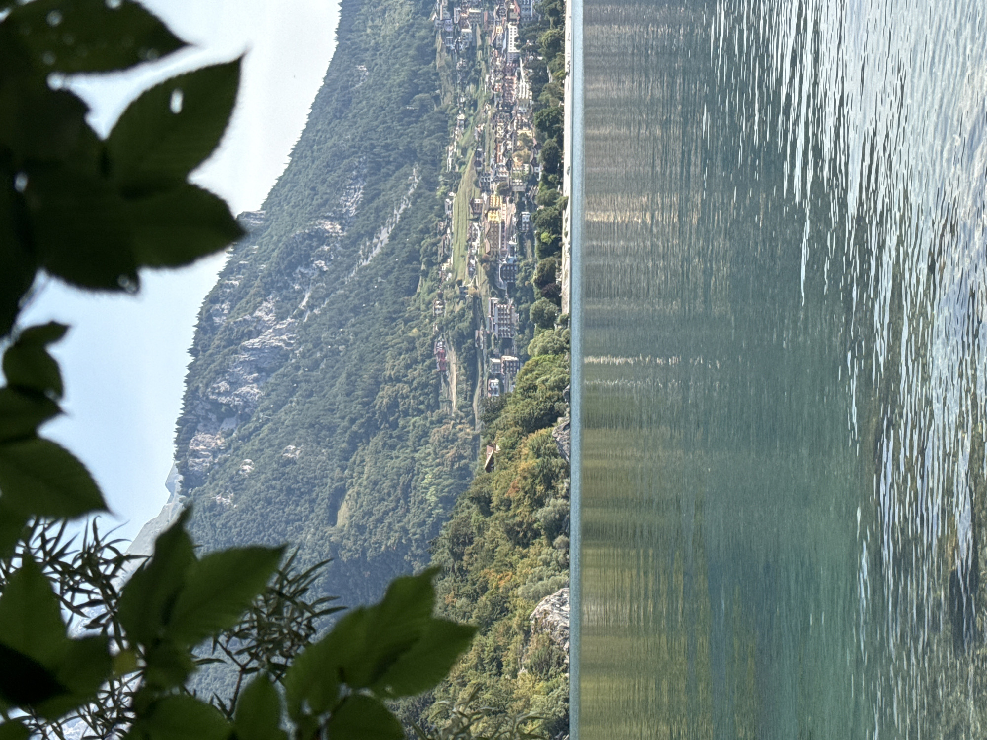 An outdoor, eye-level, vertical medium shot shows a lake in the foreground with a town built on a hill in the background. The lake is calm with light rippling on the surface. The water is a clear, light blue-green and the bottom of the lake can be seen in some areas. The town is nestled on the side of a mountain covered in trees. The buildings are a mix of residential and commercial structures, with a variety of colors and architectural styles. A few trees with green and yellow leaves are in the foreground, with their branches extending into the frame from the left. The sky is a pale blue and is clear. The time of day is daytime, and the lighting suggests a bright, sunny day. The scene is peaceful and serene. The location is identified as being in the city of Unknown, Italy, and it appears to be a resort or tourist area.