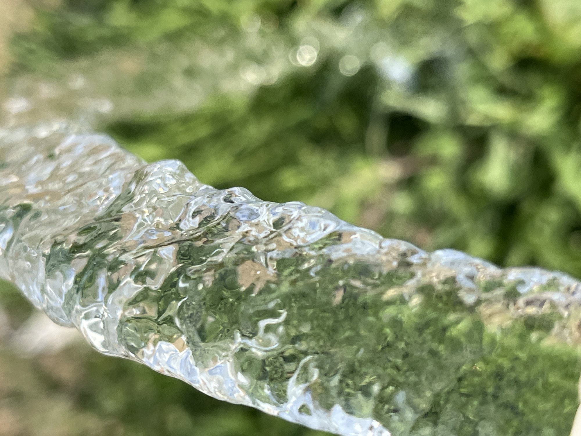 The image shows a close-up of a stream of water, likely from a hose or faucet, against a blurred green background of foliage. The water is transparent and refracts light, creating highlights and distortions. The setting appears to be outdoors, possibly in a garden or park. The specific location in Krasnyye Barrikady, Russia, is not visually discernible from the image itself. There are no people, animals, or specific identifiable man-made structures visible. The focus is on the texture and movement of the water.